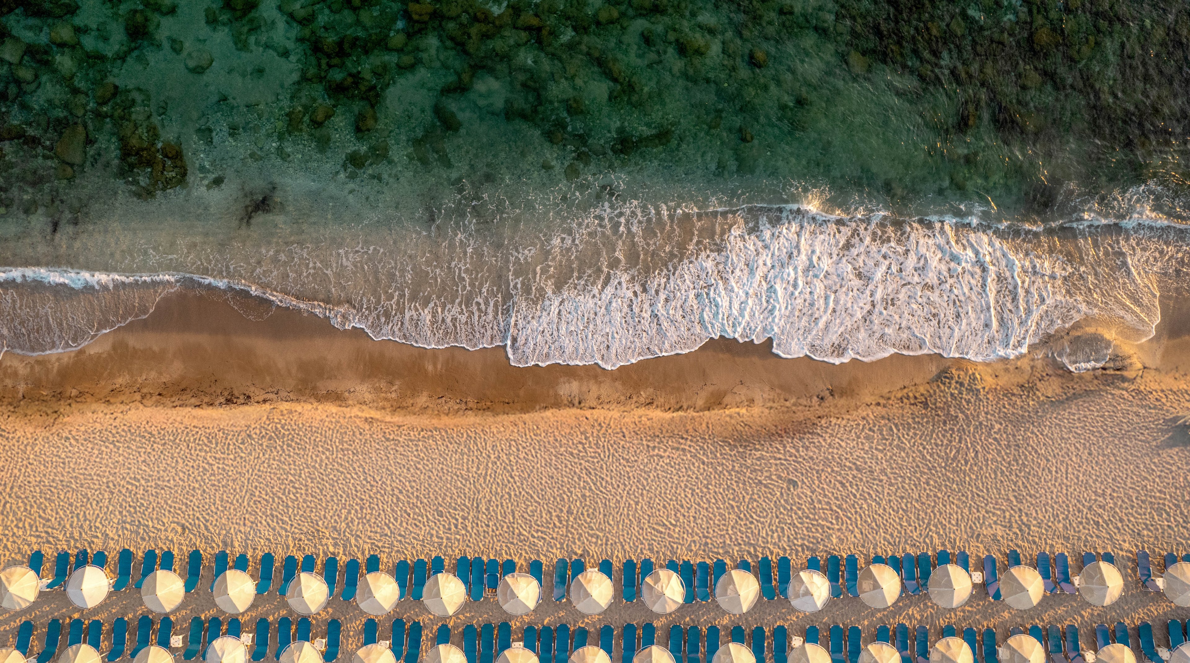 The image shows a top-down perspective of a sandy beach lined with rows of white umbrellas and blue lounge chairs. Gentle waves roll onto the shore, creating a serene coastal scene. The water transitions from deep green to lighter hues near the sand, and the umbrellas are neatly arranged in parallel lines. No visible text or numbers are present in the image.