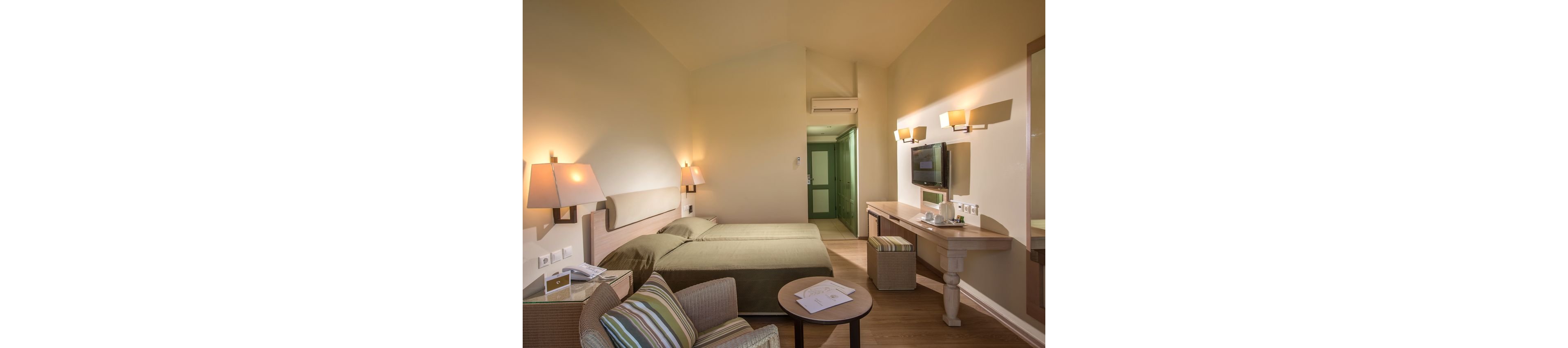 A contemporary hotel room features a double bed with neutral bedding and a small seating area. The space includes a desk with a flat-screen television mounted on the wall and a cushioned bench beneath. Warm wall sconces and table lamps create a cozy atmosphere, highlighting the light wood flooring and soft beige walls. A doorway at the back leads to an entry hall with a visible air conditioning unit above.