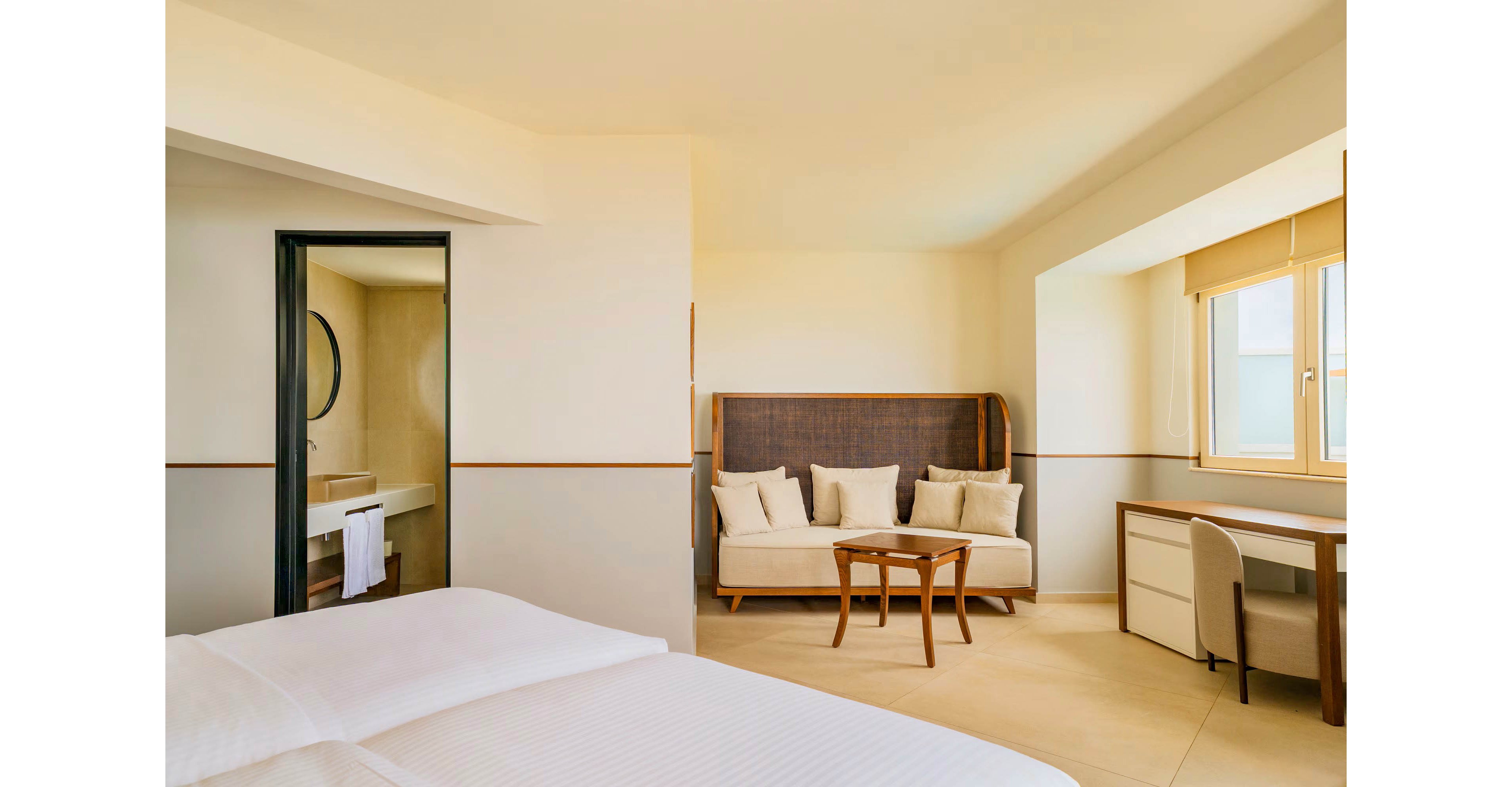 A modern hotel room features a neatly made bed in the foreground with white linens. In the background, a cozy seating area includes a cushioned sofa with multiple throw pillows and a small wooden coffee table. A desk with a chair sits beside a large window, allowing natural light to fill the space. An open doorway reveals a bathroom with a round mirror and a towel hanging near the sink, enhancing the clean and minimalist atmosphere.