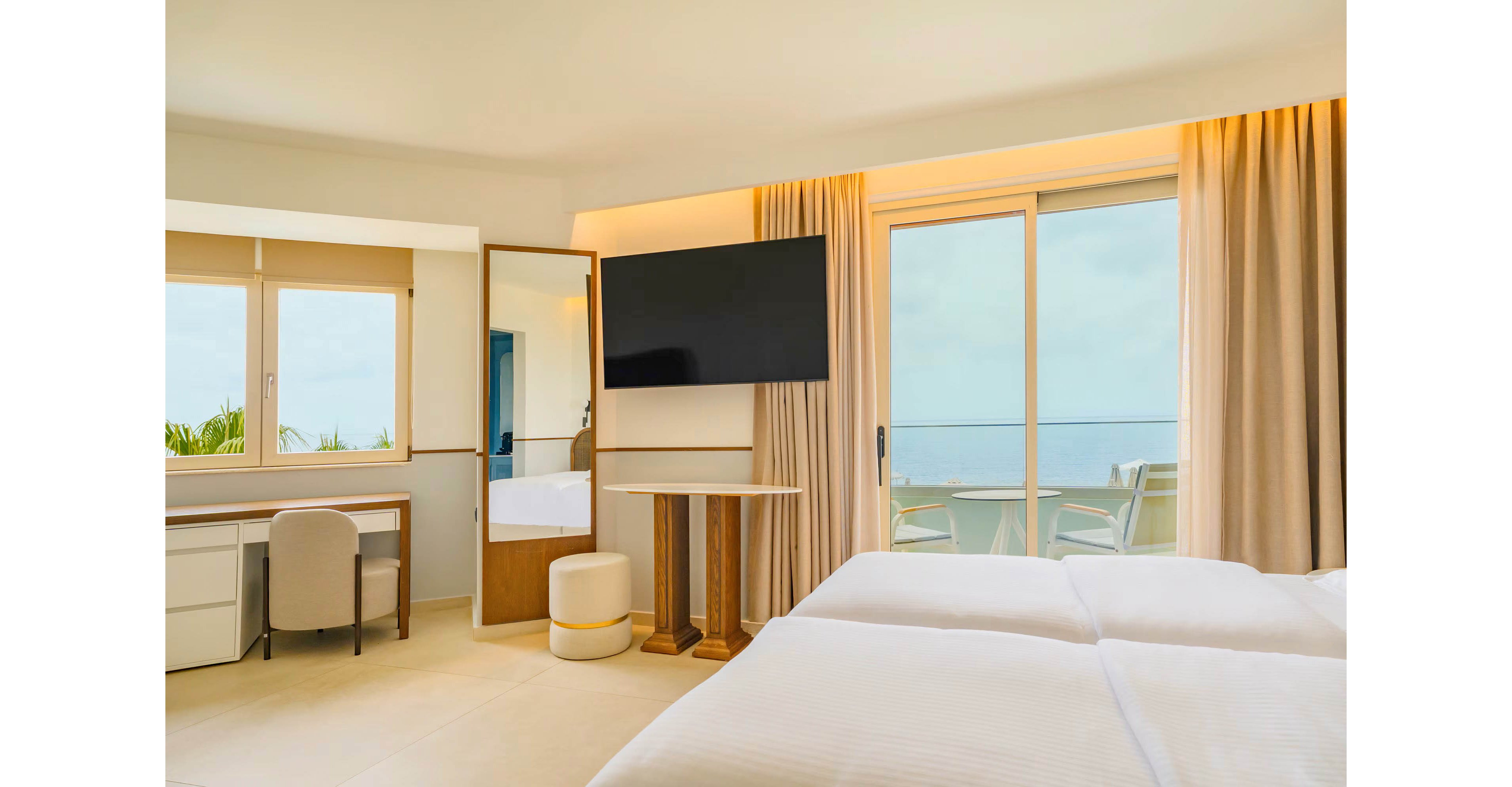 A modern hotel room features two neatly made beds facing a large flat-screen TV. Light-colored furnishings, including a desk with chair and a small round stool, create an airy, minimalist atmosphere. Floor-to-ceiling curtains frame sliding glass doors that open to a balcony with chairs overlooking the ocean. Soft natural light enters through the windows, highlighting the clean and relaxing coastal interior.