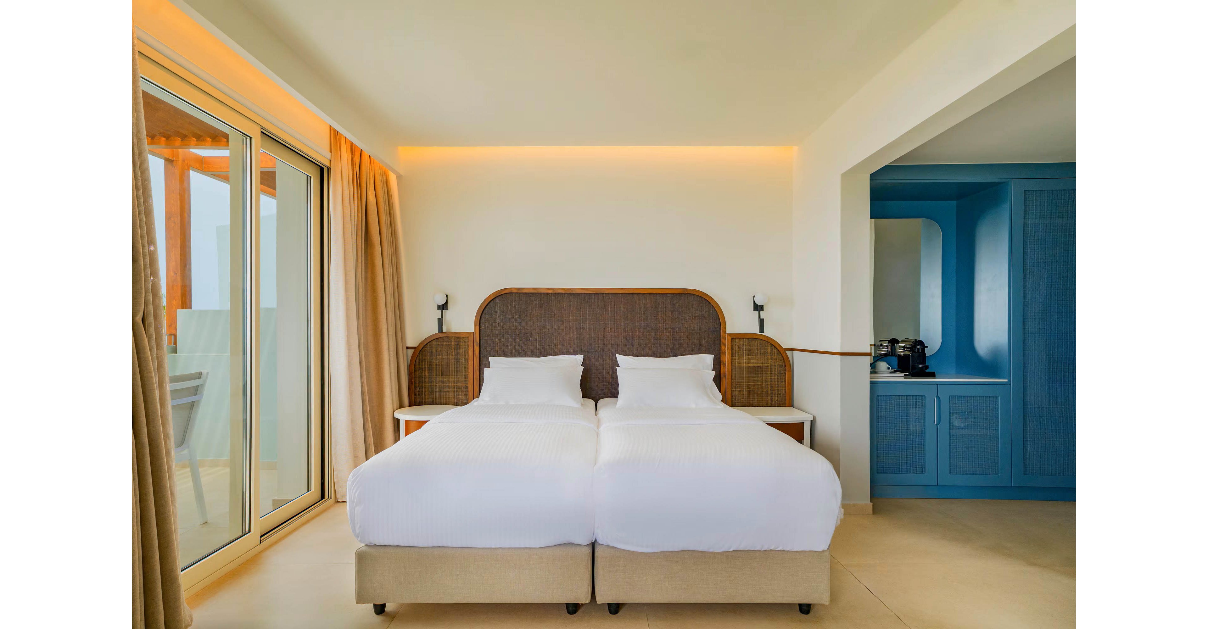 A contemporary hotel room features two neatly made twin beds with white linens and a woven headboard. Large sliding glass doors with beige curtains open to a balcony, filling the space with natural light. Warm ambient lighting runs along the wall above the bed, creating a cozy atmosphere. In the background, a blue built-in cabinet area includes a coffee machine and cups, adding convenience for guests.