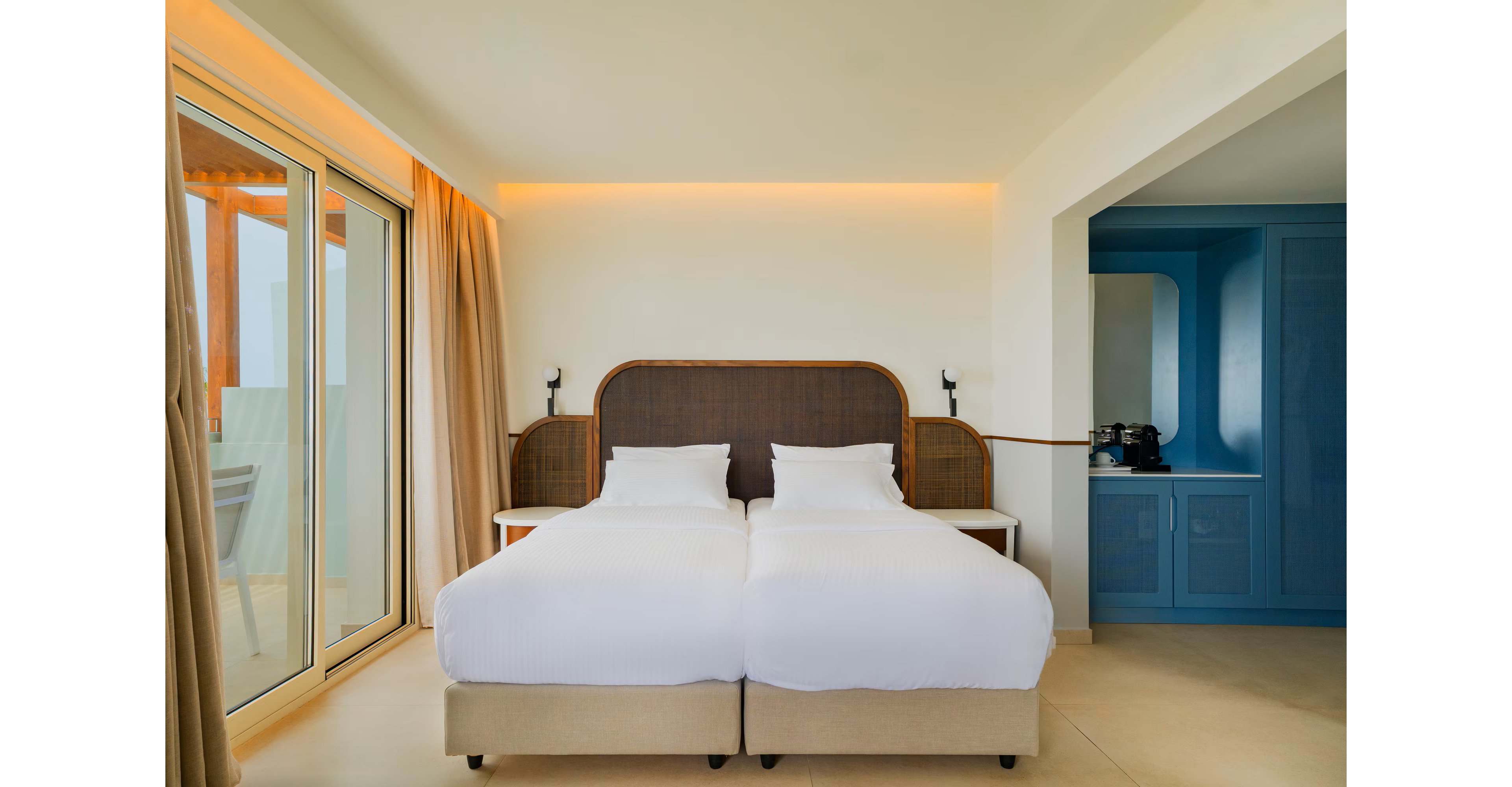 A contemporary hotel room features two neatly made twin beds with white linens and a woven headboard. Warm ambient lighting runs along the wall above the bed, creating a cozy atmosphere. Large sliding glass doors with beige curtains open to an outdoor balcony area, bringing in natural light. On the right, a blue built-in cabinet houses a coffee machine and cups, adding convenience for guests.