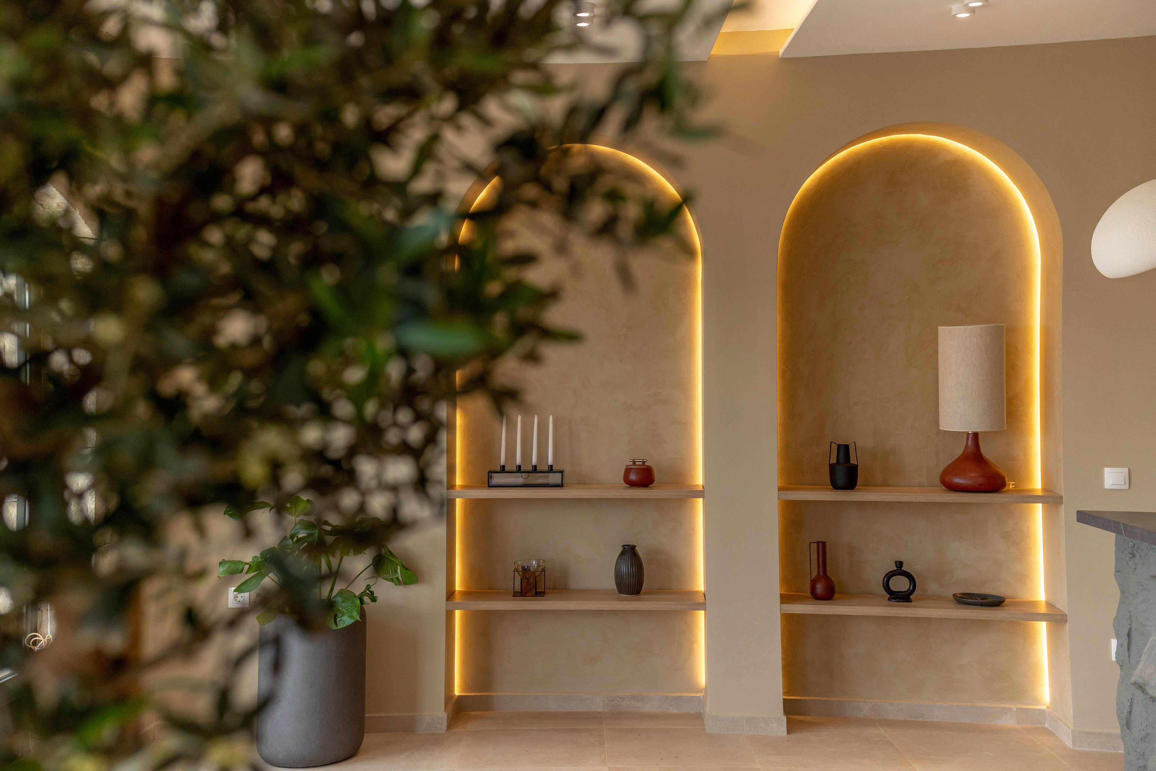 A modern interior features two arched wall alcoves with built-in shelves. Soft warm lighting outlines the arches, highlighting decorative vases, candles, and a contemporary table lamp. Neutral beige walls and light flooring create a calm, minimalist atmosphere. A blurred indoor plant in the foreground adds depth and a natural touch to the scene.