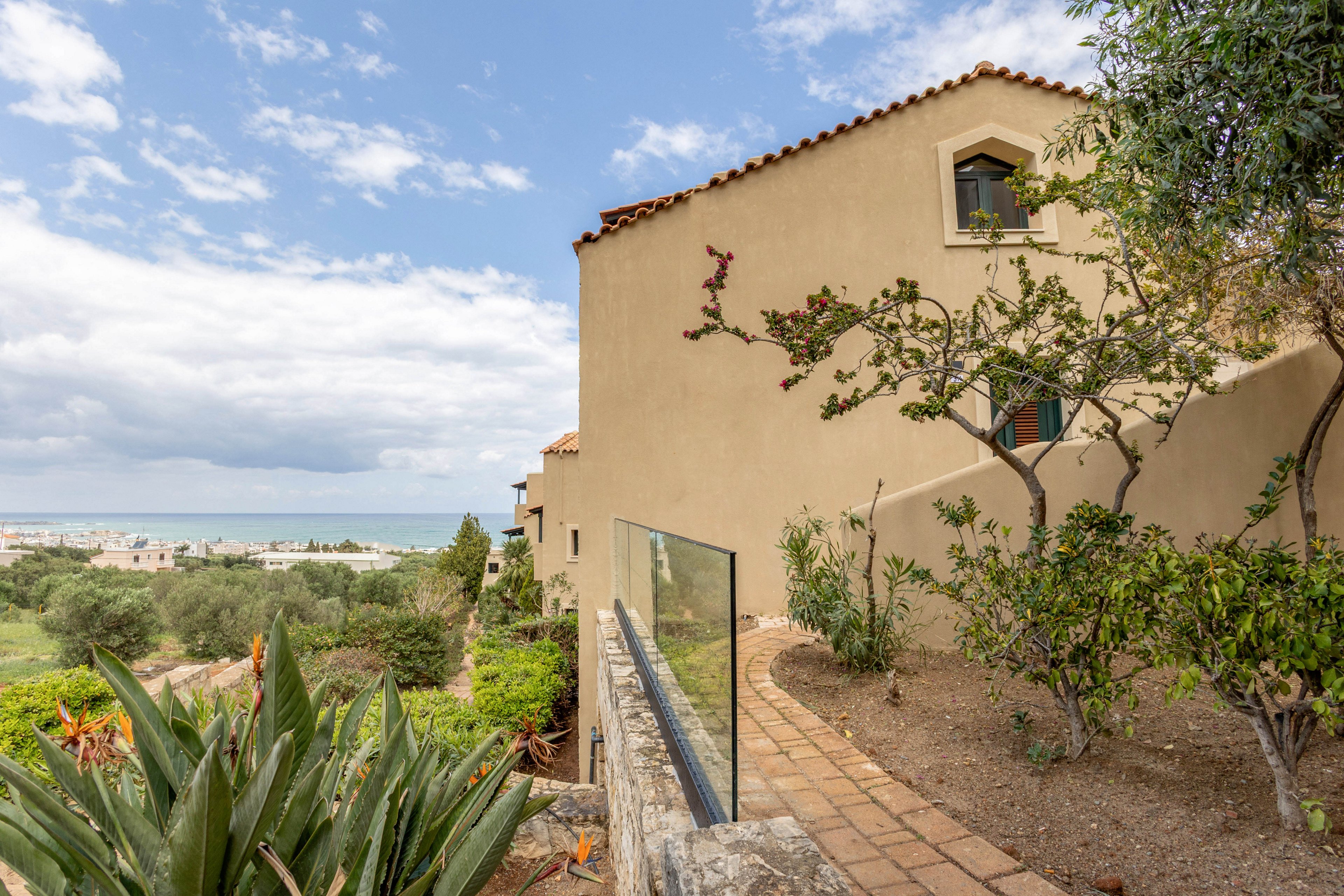 A Mediterranean-style villa sits on a hillside overlooking a distant coastal town and the sea. The building features stucco walls, terracotta roof tiles, and a narrow brick pathway bordered by a glass railing. Lush greenery, including succulents and small trees, surrounds the house and garden area. Soft clouds fill the sky, creating a calm and relaxed atmosphere in this scenic landscape.