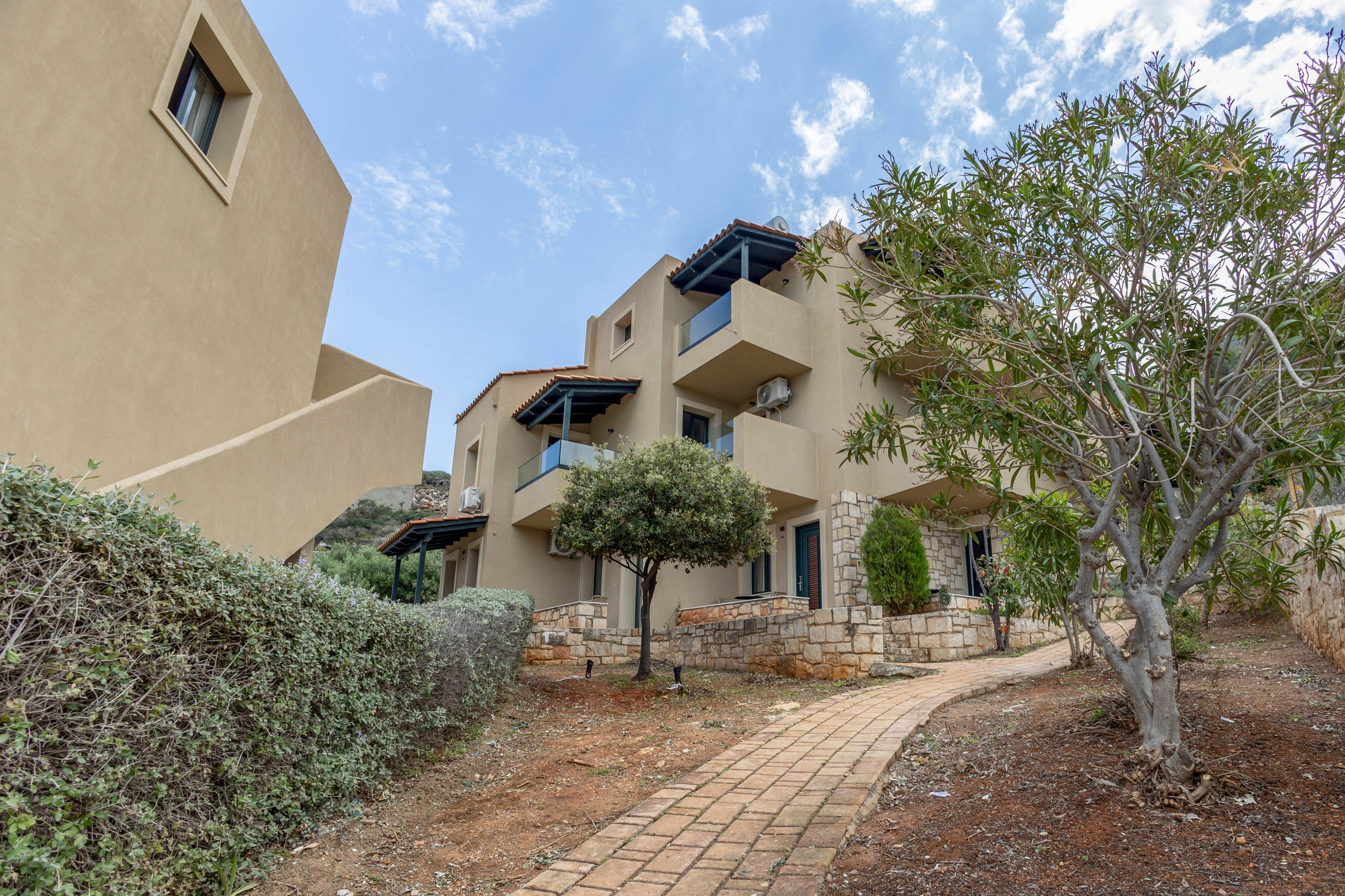 A beige, multi-story apartment building stands in a landscaped outdoor setting. A curved brick walkway leads past shrubs and small trees toward the entrance and balconies. The architecture features simple geometric forms, flat roofs, and small terraces with railings. The scene appears calm and residential under a partly cloudy sky.