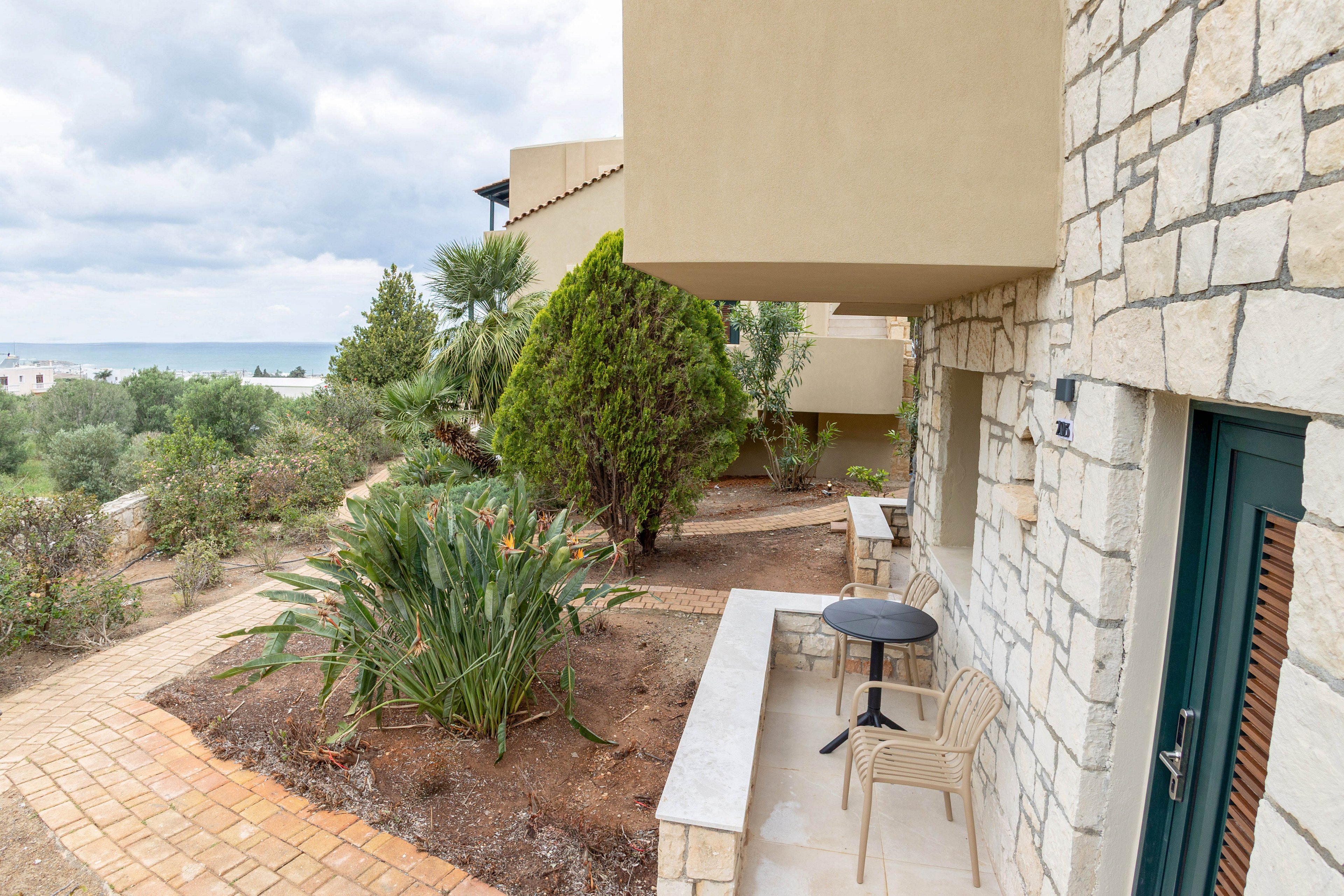 A small stone terrace with simple outdoor chairs and a round table overlooks a landscaped garden. The building features light stone walls and beige stucco, giving a Mediterranean feel. Pathways and shrubs lead toward the distant coastline and sea under a cloudy sky. The scene suggests a quiet holiday accommodation or resort setting with natural surroundings.