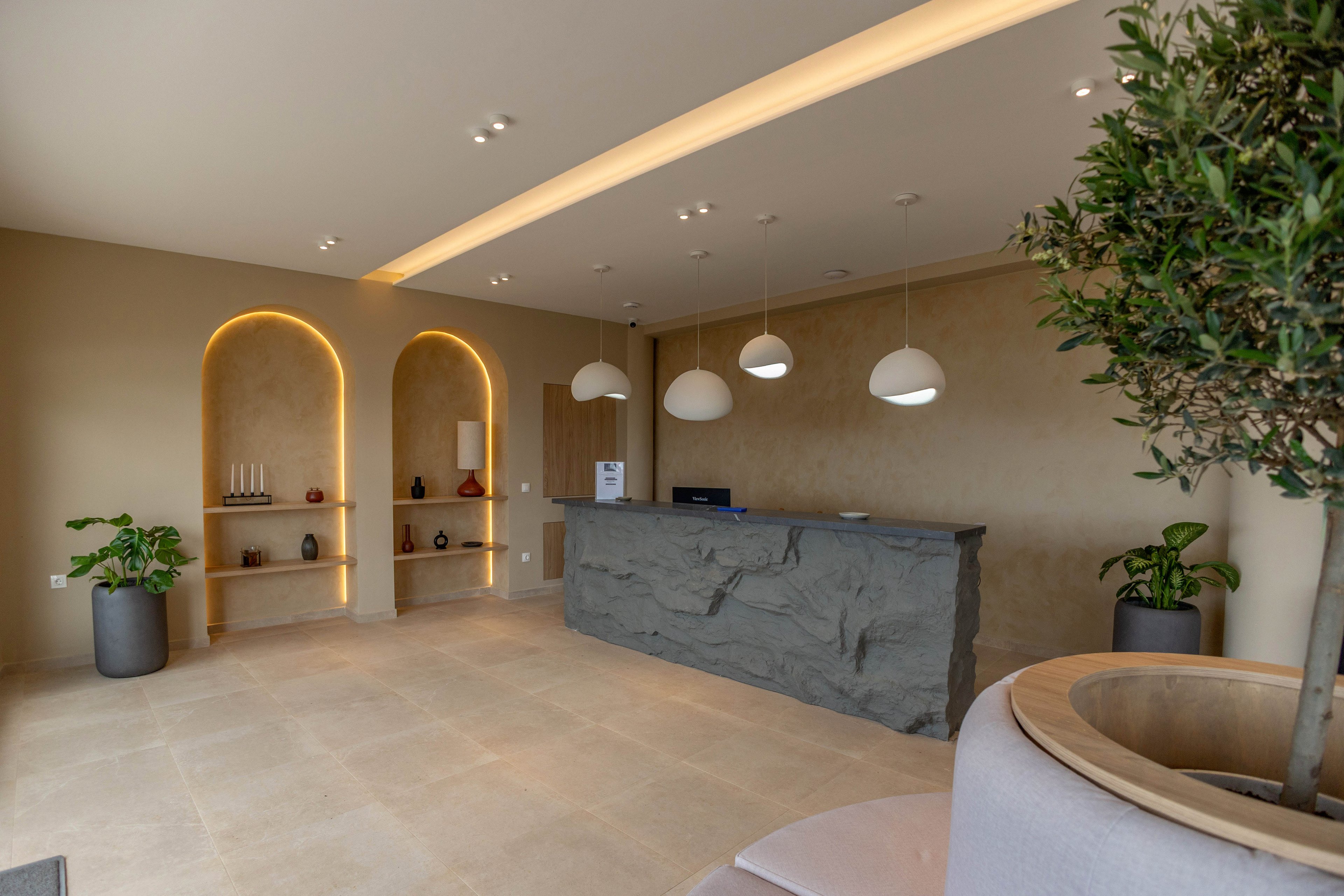 A contemporary reception desk stands in a minimalist lobby with warm beige walls and stone-like finishes. Built-in arched wall niches with soft backlighting display decorative vases and objects. Three pendant lights hang above the counter, while potted plants and a large indoor tree add greenery to the space. The overall atmosphere is calm, welcoming, and modern.
