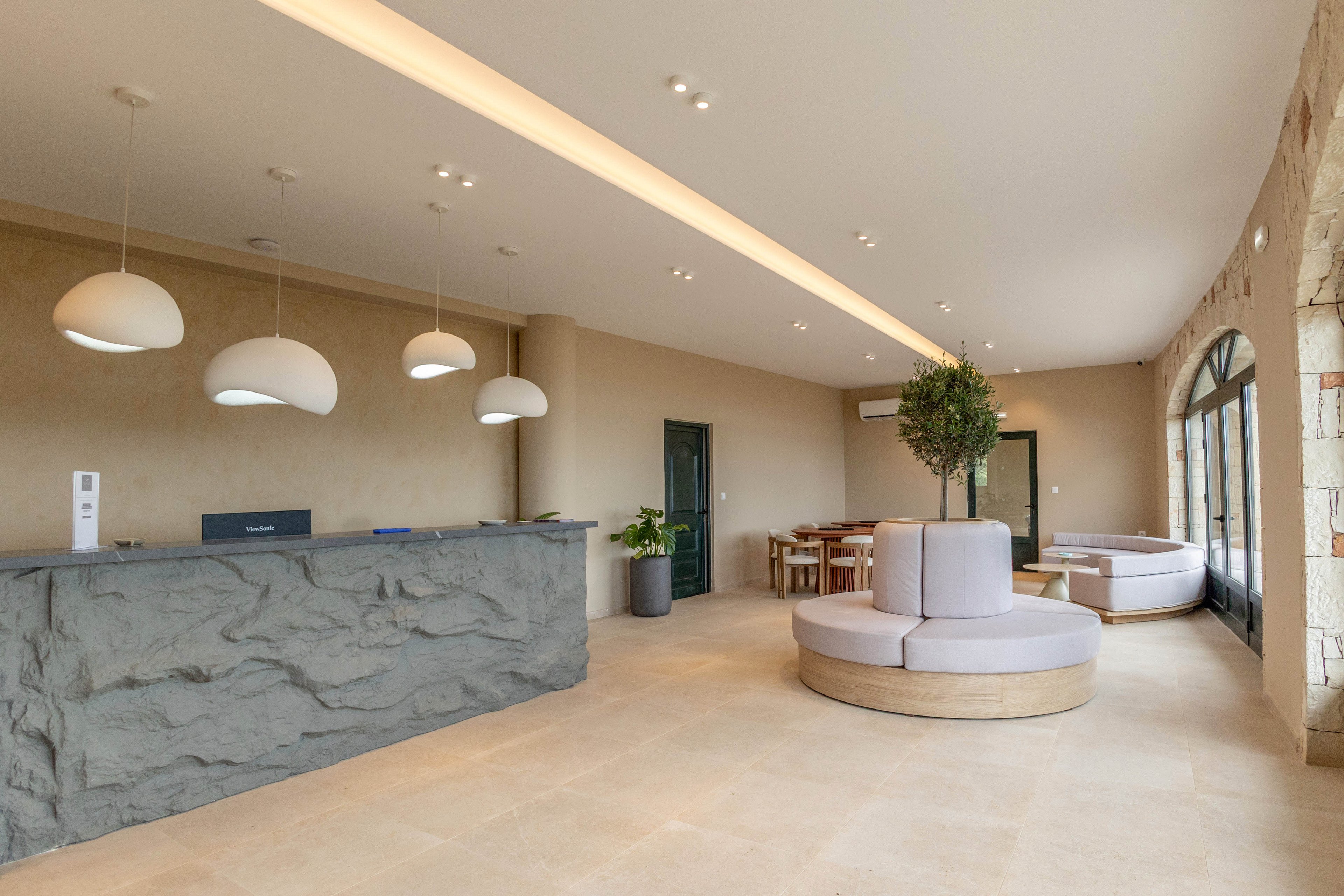 A contemporary hotel lobby features a large stone-textured reception desk on the left side. Minimalist pendant lights hang above the counter, illuminating the spacious interior. In the center, a round upholstered seating area and potted plants add comfort and greenery. Large windows and glass doors on the right bring in natural light, highlighting the neutral color palette and clean architectural lines.