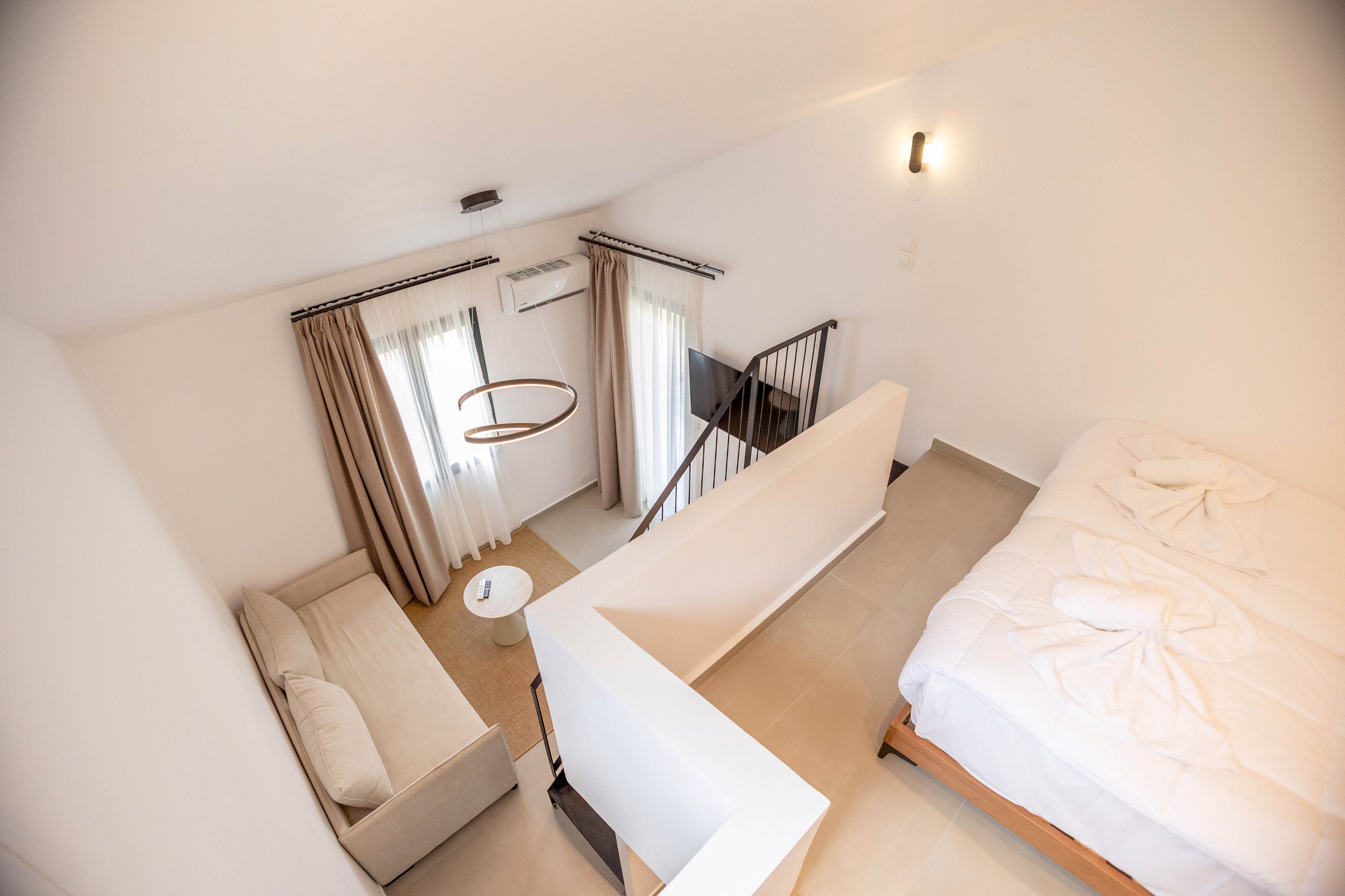 A compact loft-style bedroom features a neatly made bed beside a low partition wall. Below the loft, a small sitting area includes a light-colored sofa, round side table, and floor-to-ceiling curtains framing tall windows. A modern circular ceiling light and wall sconce provide warm illumination in the otherwise white interior. A staircase with a black metal railing connects the upper sleeping area to the lower level, emphasizing the open-plan design.