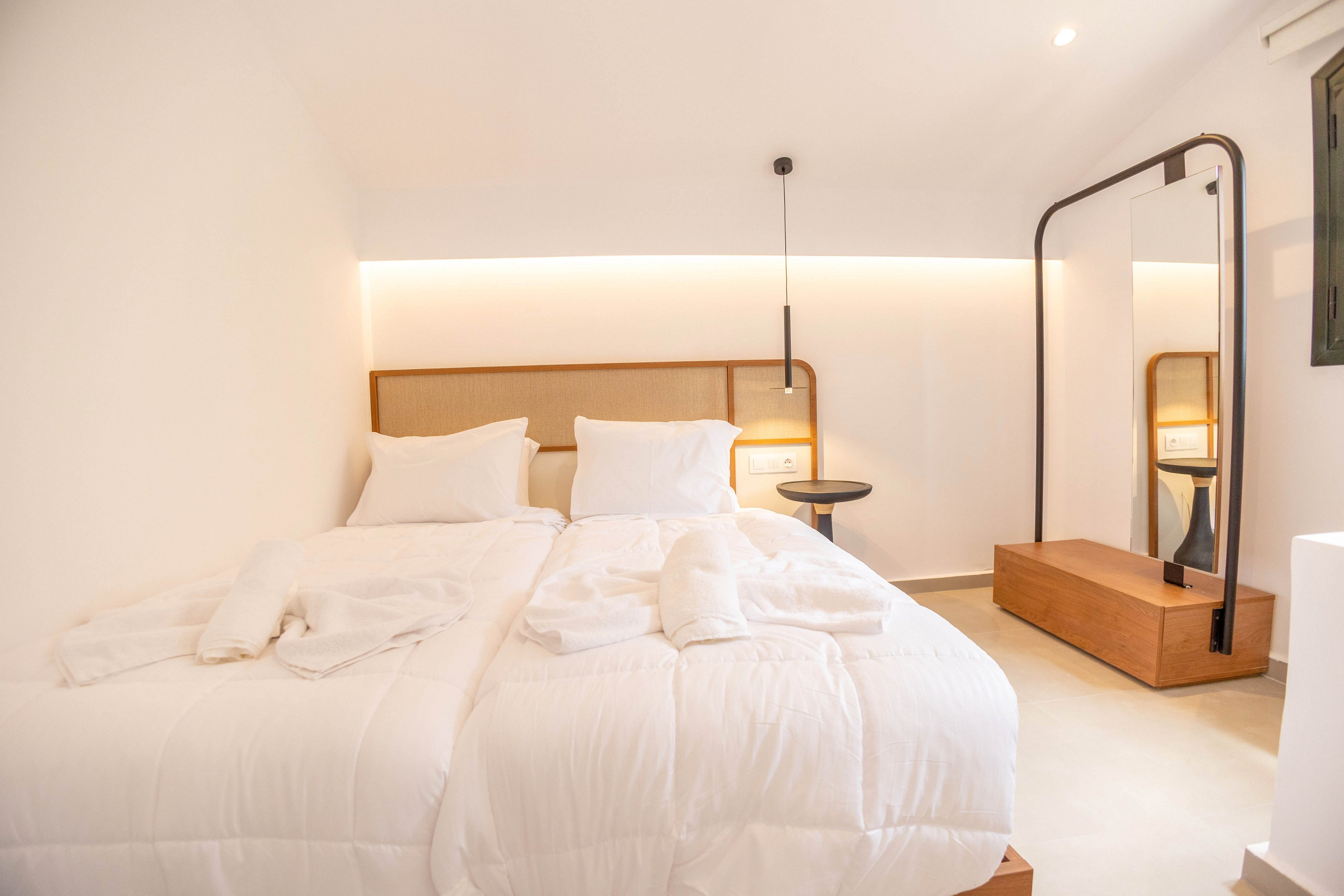 A bright minimalist bedroom features a neatly made double bed with fluffy white bedding and pillows. Soft indirect lighting runs along the wall behind the headboard, creating a warm and inviting atmosphere. A modern floor mirror with a wooden base and a small bedside table with a lamp complete the simple decor. The room appears clean, contemporary, and ready for guests.