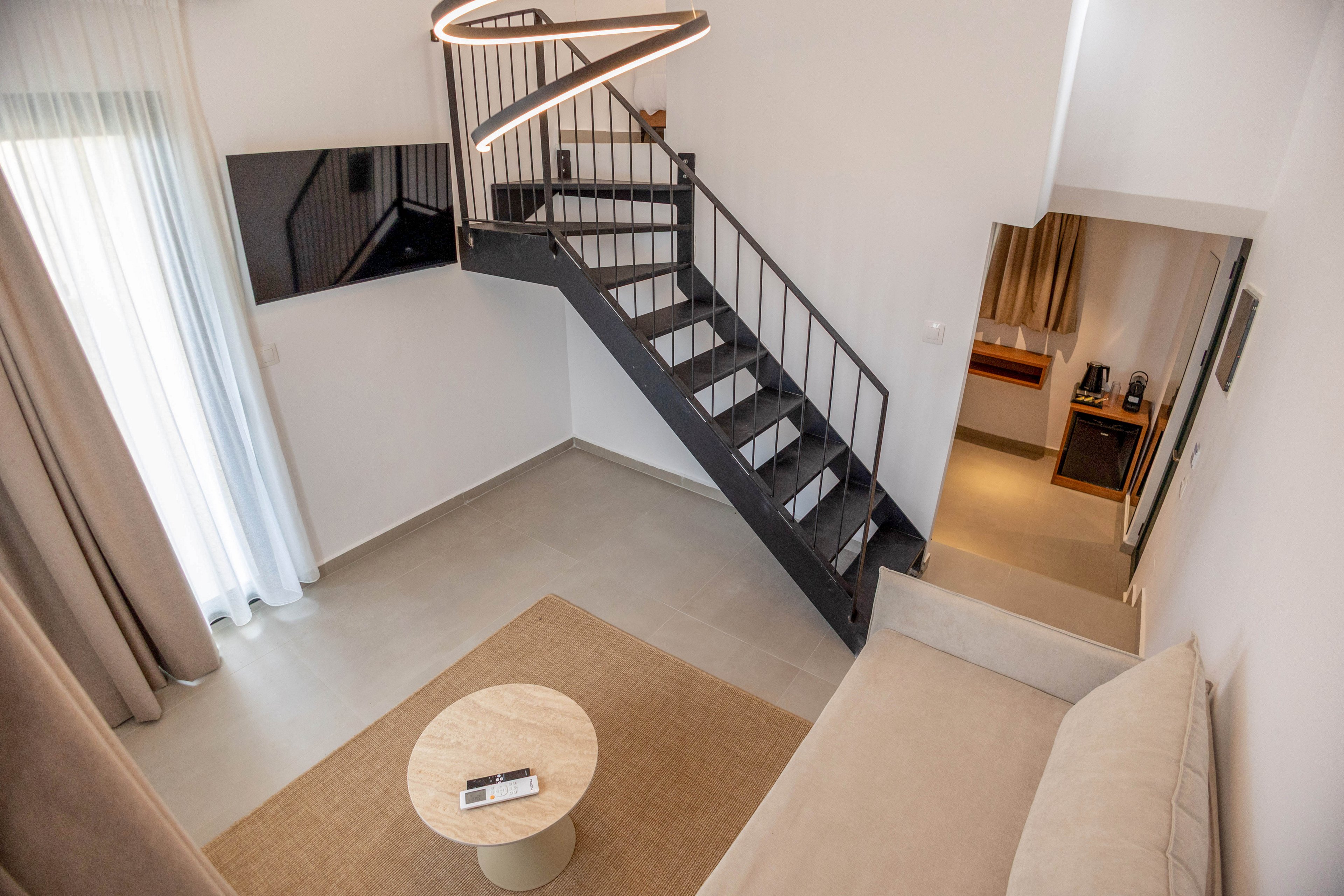 A contemporary loft-style living room features a beige sofa and a small round coffee table viewed from above. A sleek black metal staircase leads to an upper level, adding an architectural focal point to the space. Large curtains filter natural light from a tall window, while a wall-mounted flat-screen TV hangs opposite the seating area. In the background, a small workspace or kitchenette area is partially visible, enhancing the sense of a compact yet stylish interior.
