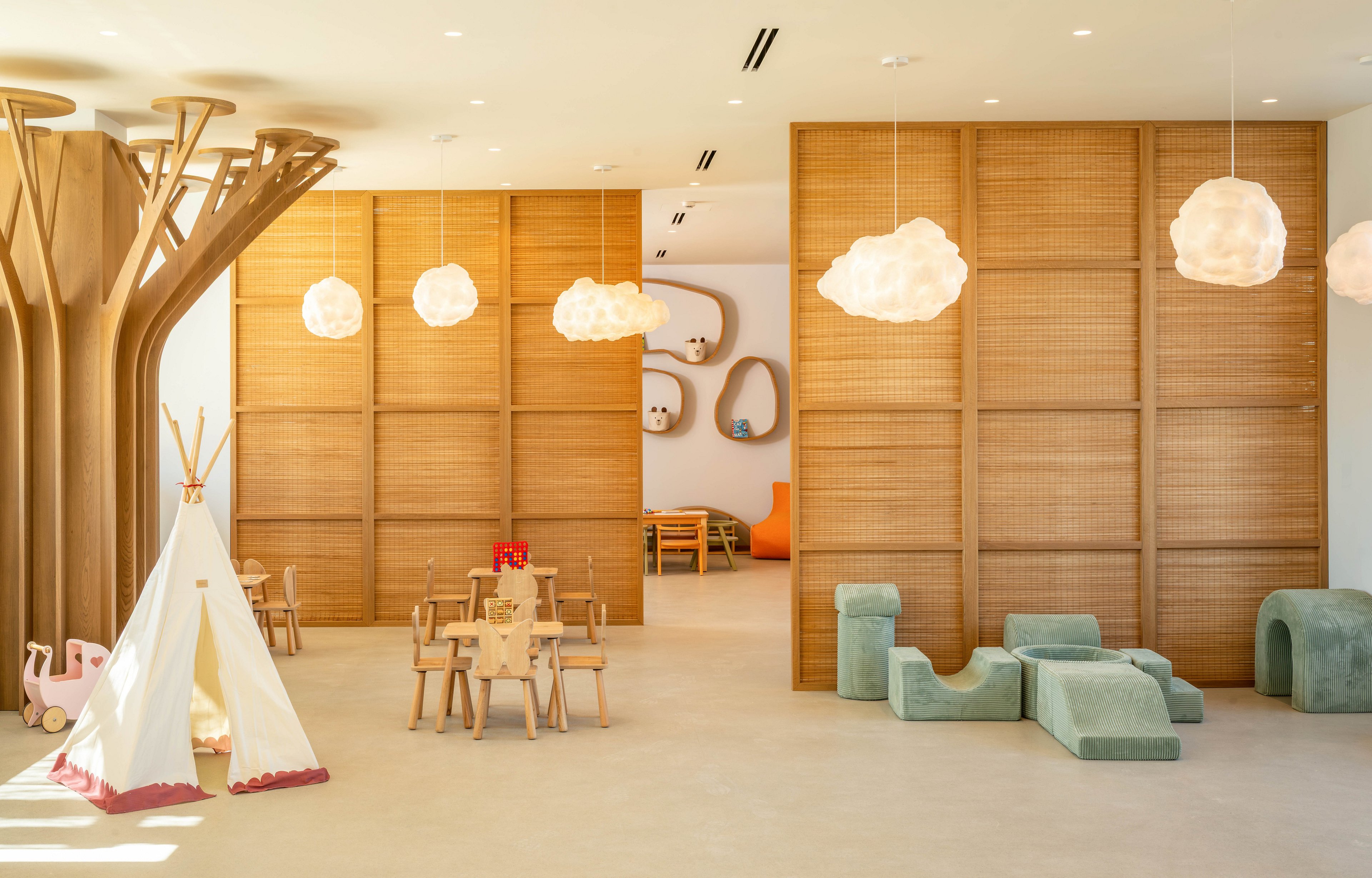 A bright, modern children's playroom features light wooden furniture and sculptural tree-like columns. Soft cloud-shaped pendant lights hang from the ceiling, illuminating a teepee tent, small tables, and chairs. Curved foam play blocks and built-in wall nooks add playful shapes and textures to the space. The neutral palette of beige, tan, and soft green creates a calm, inviting environment for kids.