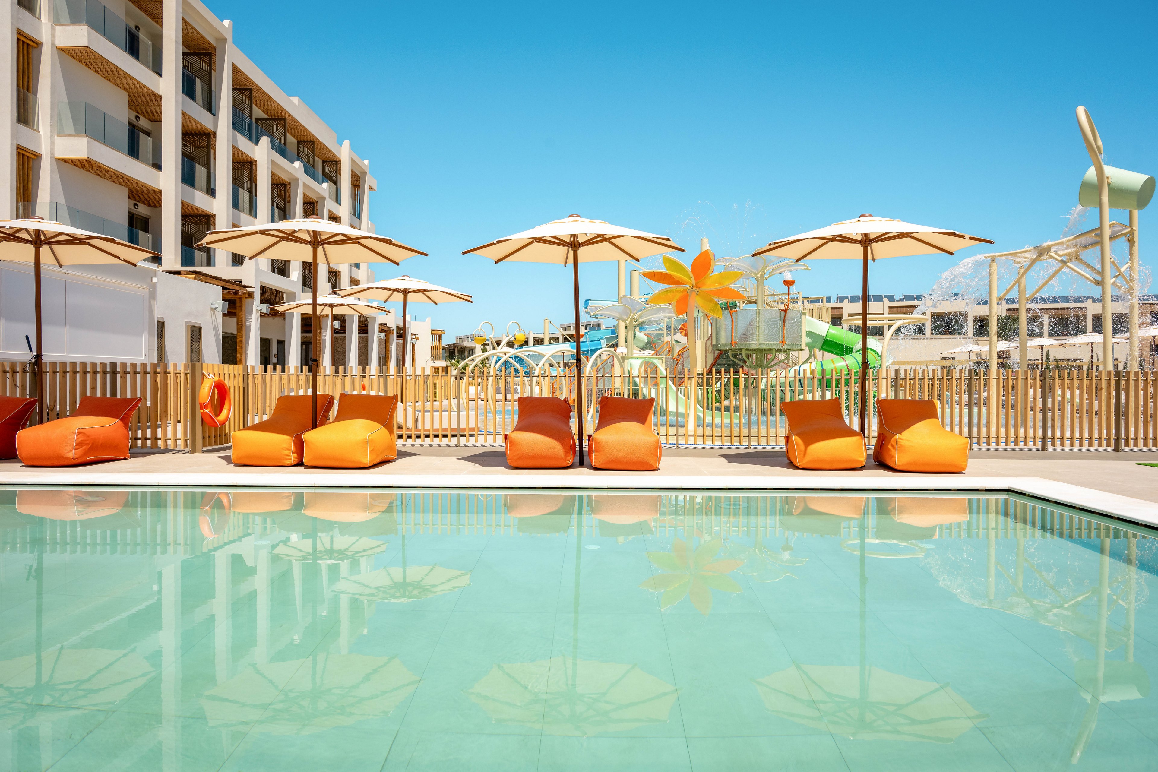 A tranquil resort swimming pool reflects the clear blue sky and surrounding structures. Bright orange and yellow loungers sit neatly in a row beneath white umbrellas along the poolside. In the background, a wooden fence separates the pool from a playful water park area with slides and large flower-shaped features. The scene suggests a warm, sunny day ideal for relaxation and family vacations.