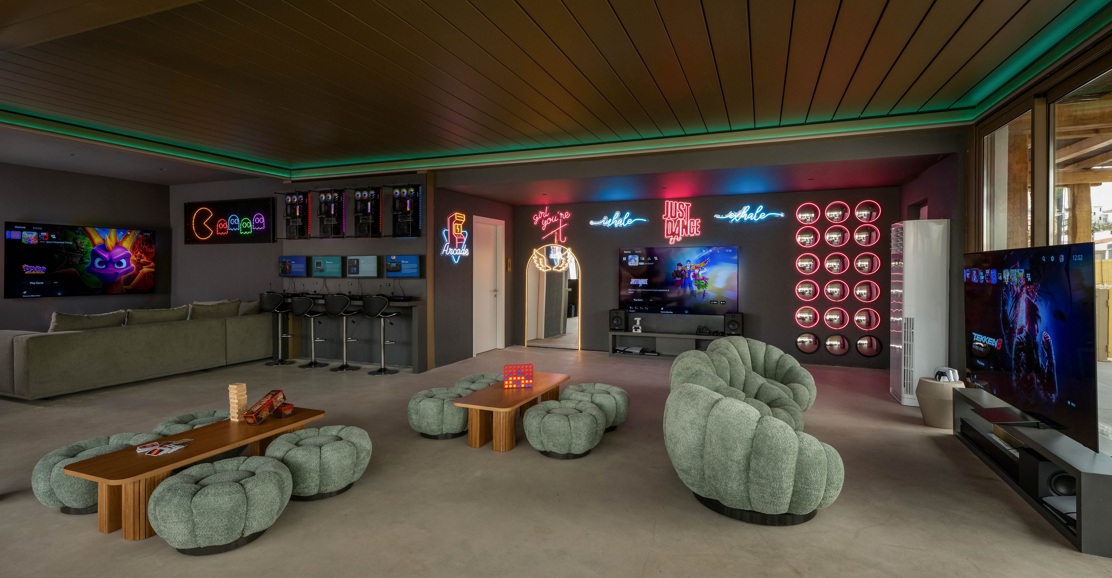 Spacious contemporary lounge features multiple large gaming screens and plush modular seating. A row of gaming PCs with monitors lines the back wall, while consoles and big-screen TVs are mounted around the room. Colorful neon signs and LED accent lights create a vibrant, futuristic atmosphere. Low wooden coffee tables with controllers and accessories emphasize the entertainment and gaming focus of the space.