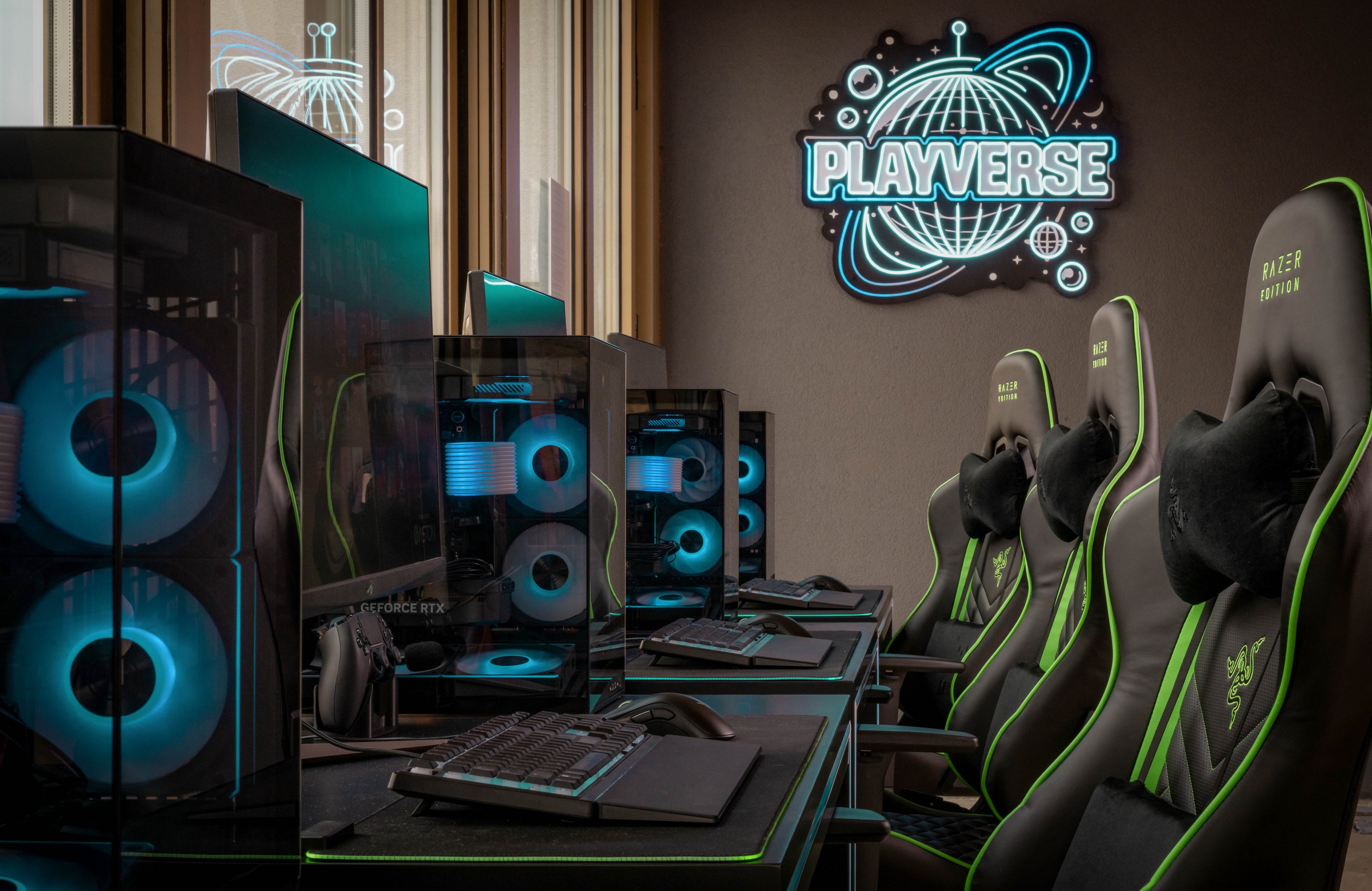 Row of high-end gaming PCs with glowing blue fans is arranged in a sleek esports station. Ergonomic gaming chairs with green accents line up beside RGB-lit keyboards and mice. A neon-style wall sign reading "PLAYVERSE" with a stylized planet graphic illuminates the room. The overall atmosphere is modern, immersive, and designed for competitive or team gaming sessions.