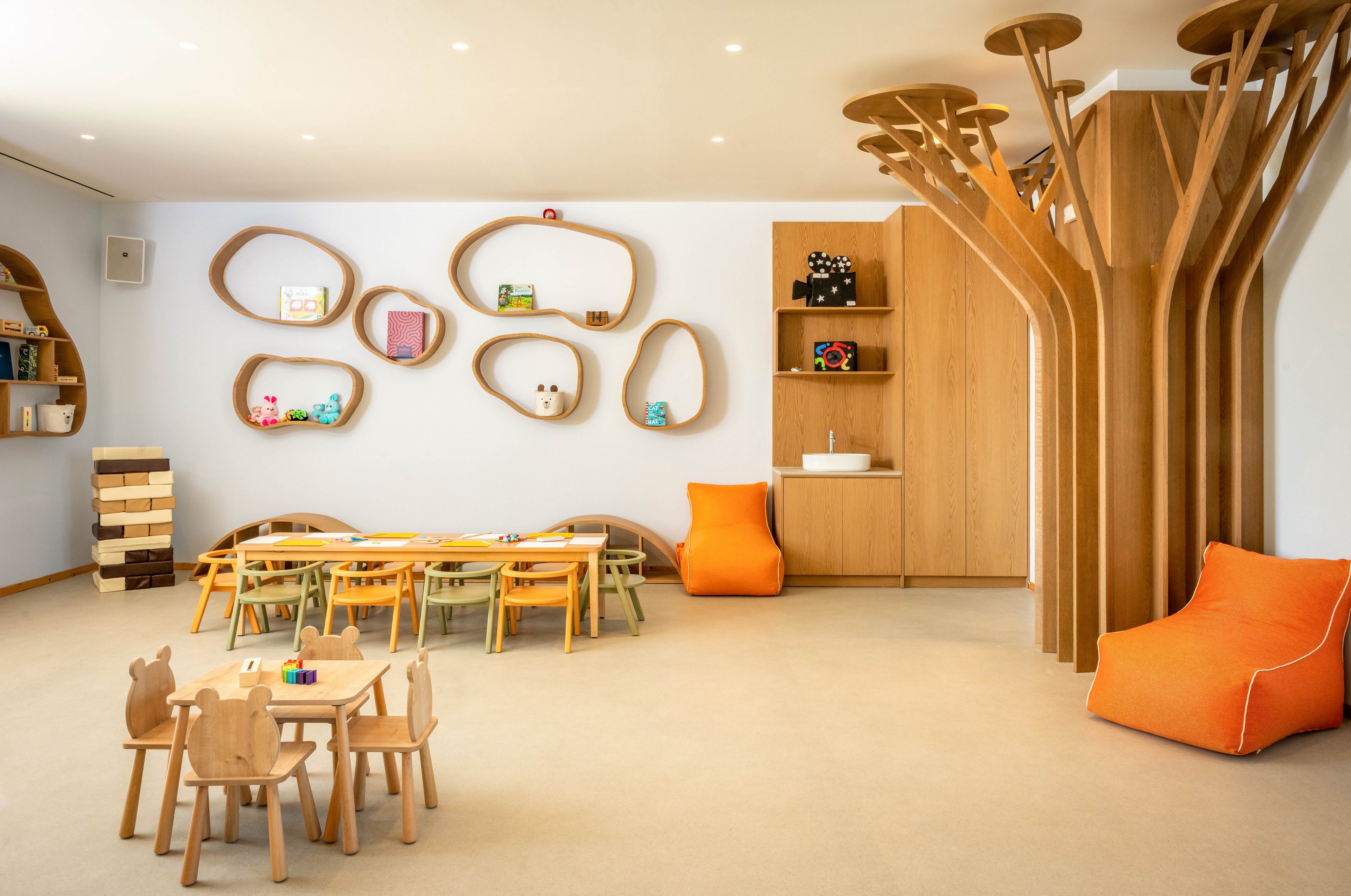 Contemporary children's playroom featuring light wooden tables and small chairs is shown in a bright, spacious interior. The room includes whimsical tree-shaped wooden structures and organic wall shelves displaying toys and books. Soft orange lounge chairs add a pop of color against the neutral walls and flooring. Overall, the space feels playful, minimalist, and inviting for kids.