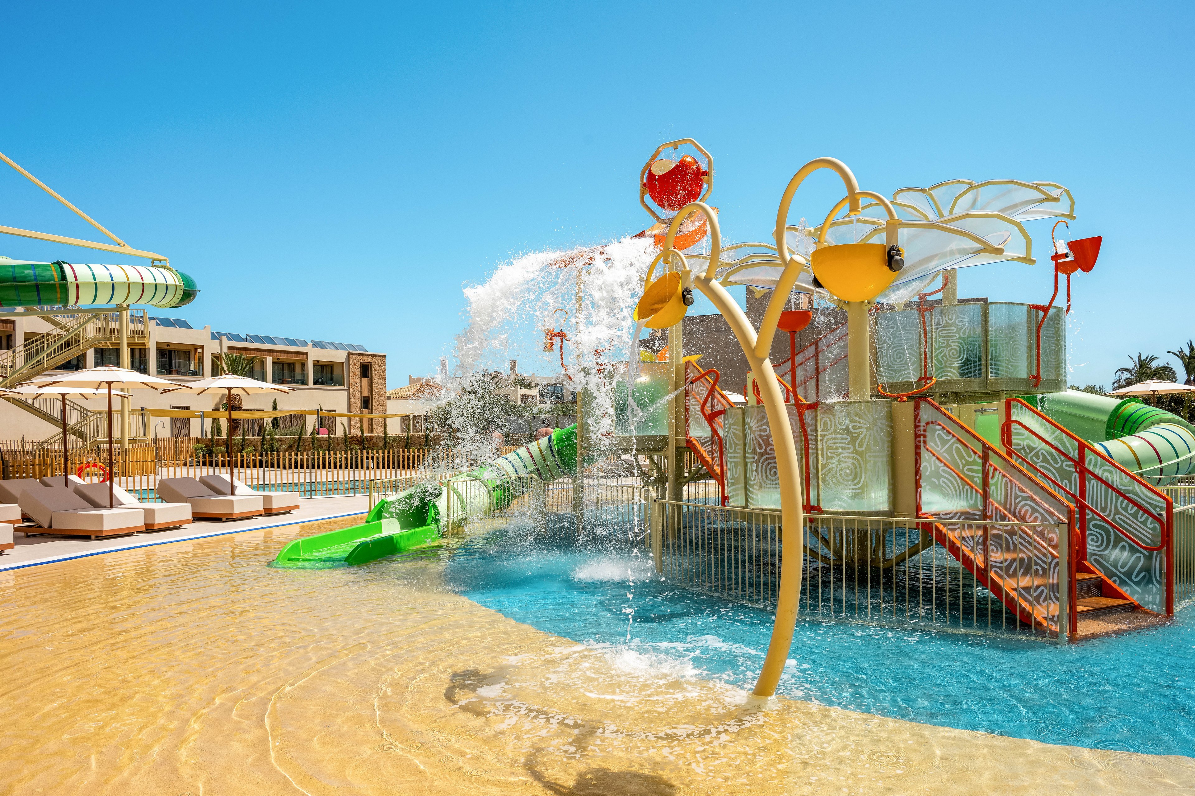A vibrant water playground features slides, splash buckets, and climbing structures in a shallow pool. The scene is set at a sunny resort with lounge chairs and modern buildings in the background. Water pours from overhead buckets, creating playful splashes and movement. Bright colors and clear blue water evoke a fun, family-friendly vacation atmosphere.