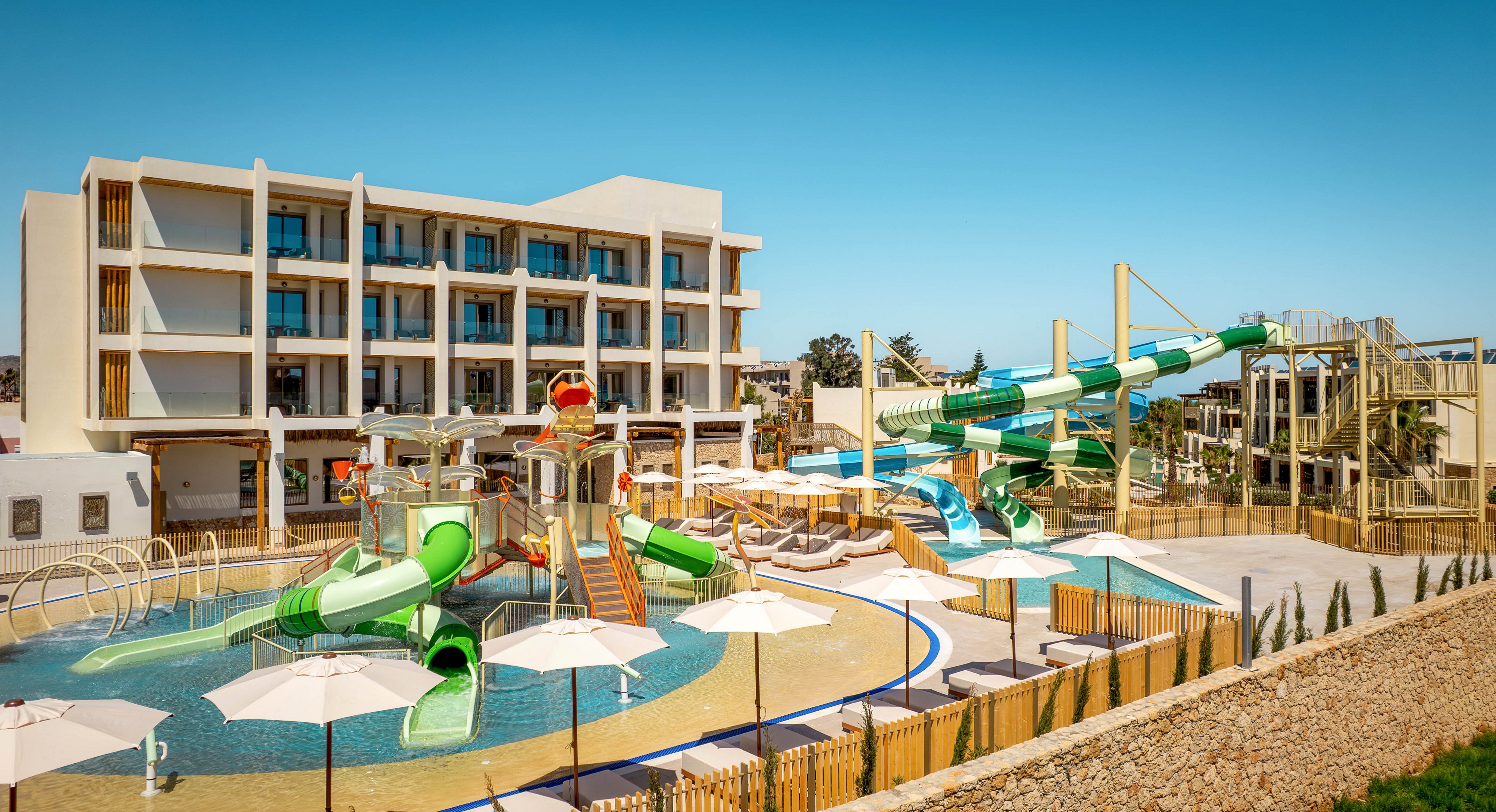 A modern multi-story resort hotel overlooks a colorful outdoor water park. Several twisting water slides and splash features lead into a shallow pool area designed for families and children. White sun umbrellas and lounge chairs line the poolside, separated by wooden fencing and landscaping. The bright blue sky and clear weather create a cheerful, summery vacation atmosphere.