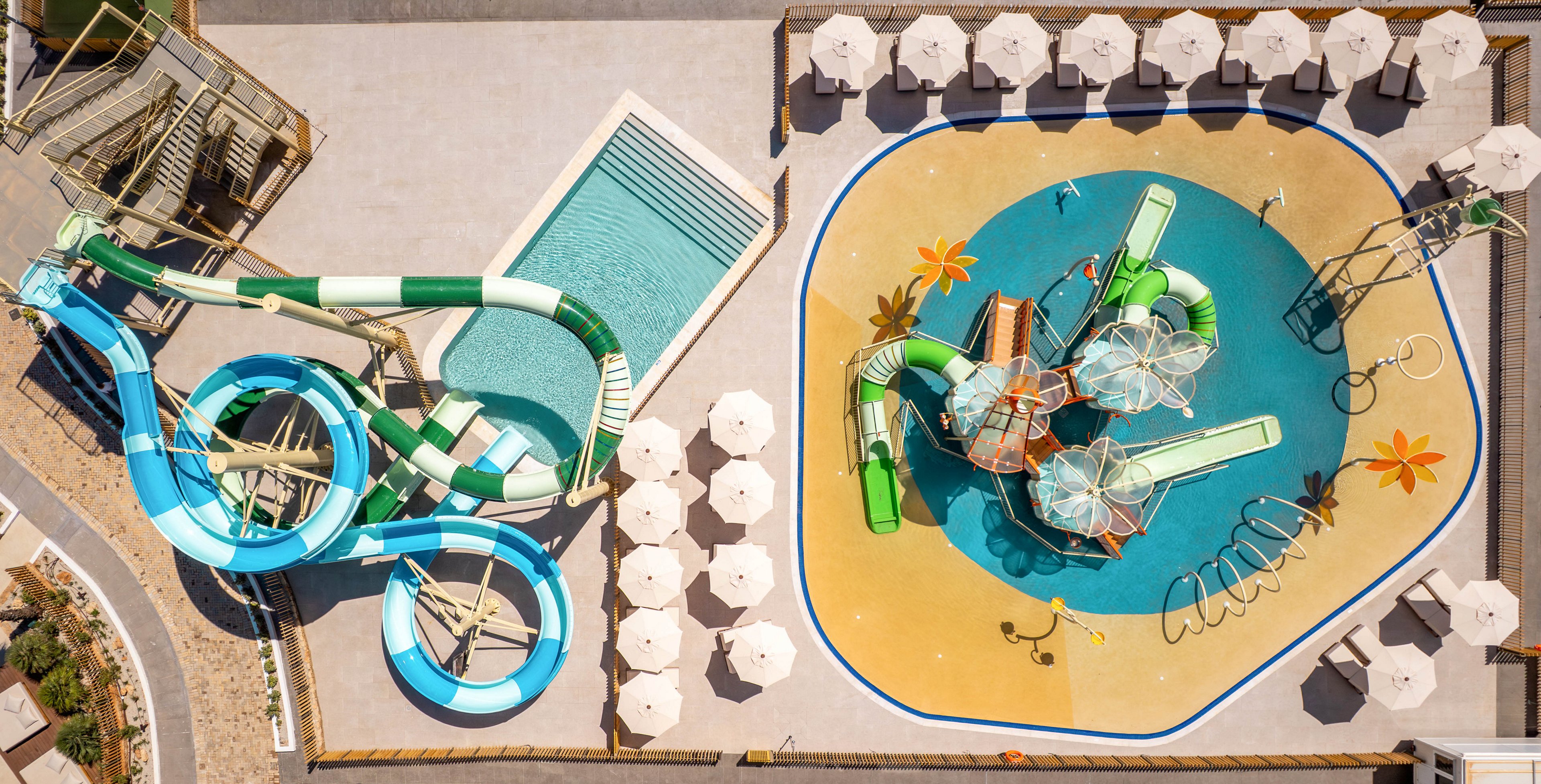 A large outdoor waterpark is photographed from above, showing twisting waterslides and splash play structures. On one side, multiple enclosed and open slides curve into a rectangular swimming pool. On the other side, a shallow splash pad features small slides, water buckets, and spray elements on a bright blue and yellow surface. Rows of white sun umbrellas and lounge chairs surround the attractions, suggesting a sunny vacation resort setting.