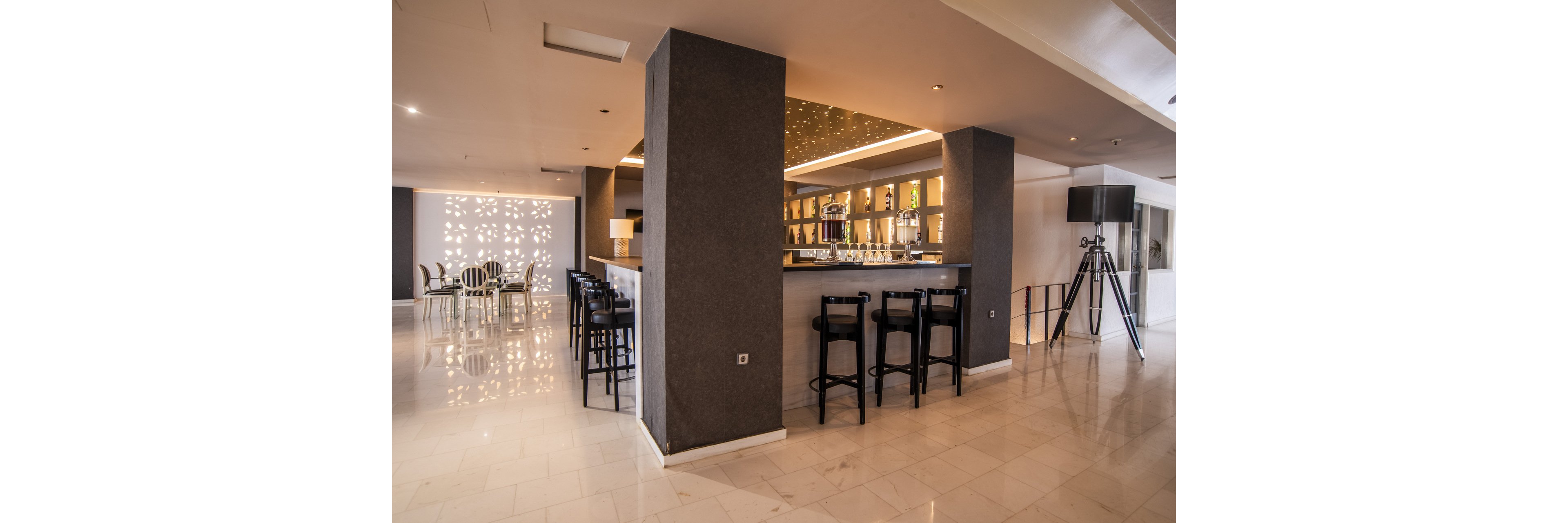 A contemporary hotel bar features a sleek counter with high stools and illuminated shelves of bottles behind it. The space is open and bright, with polished light-colored floors and neutral-toned walls. To the left, a separate seating area with tables and chairs is highlighted by decorative wall cutouts that cast patterned light. The overall atmosphere is clean, modern, and inviting, suitable for socializing or relaxing.