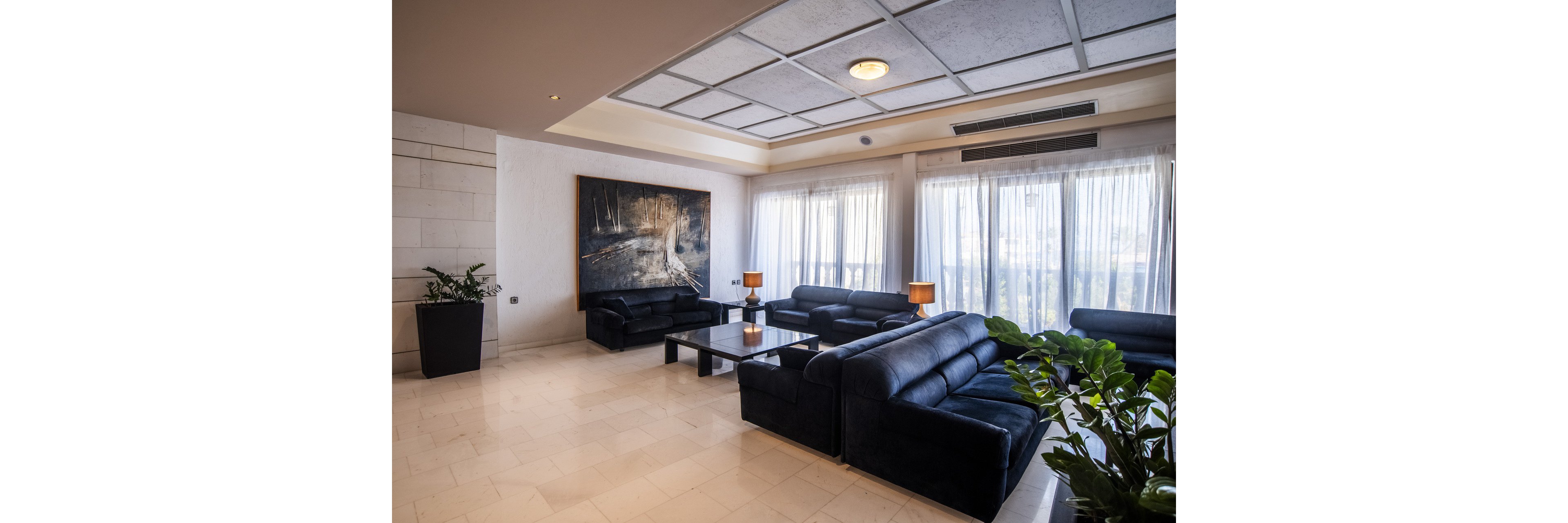 A contemporary lounge area features several black leather sofas arranged around low glass coffee tables. Large floor-to-ceiling windows with sheer curtains fill the room with natural light. Warm table lamps sit on side tables, adding a cozy glow to the modern interior. Potted plants and a large wall painting contribute to the stylish, welcoming atmosphere.