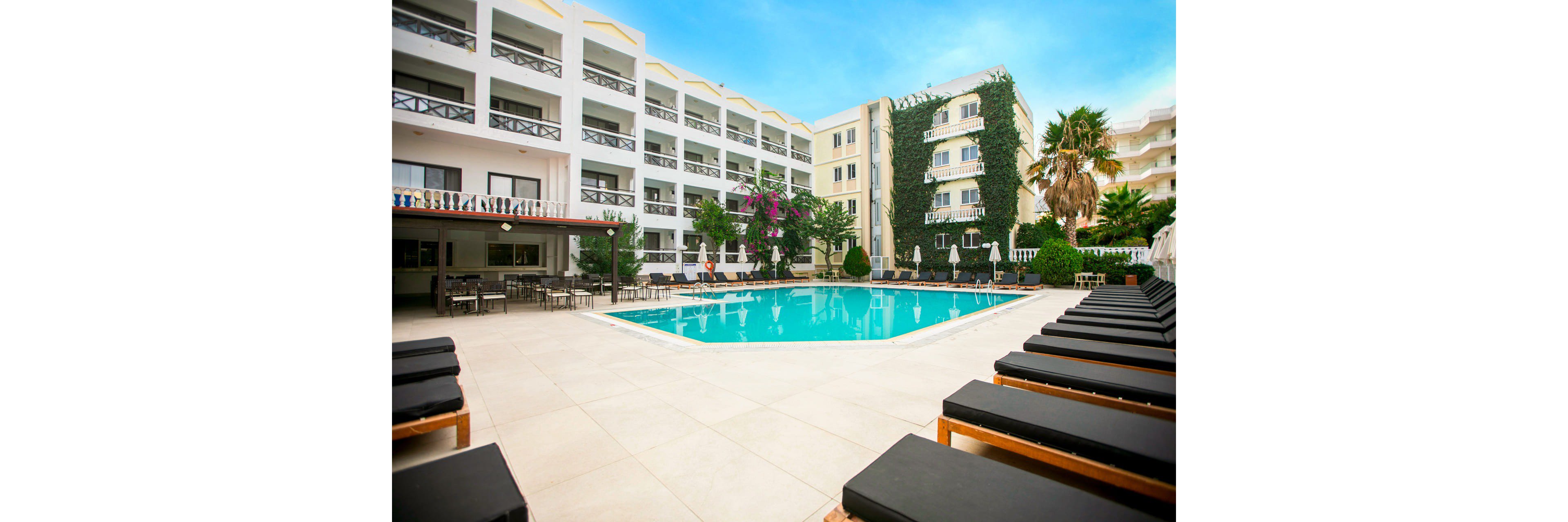 A modern multi-story hotel surrounds a central outdoor swimming pool. Black cushioned lounge chairs line the tiled pool deck, creating a relaxing sunbathing area. The buildings feature balconies, large windows, and a wall covered with green ivy and flowering plants. Clear blue sky and calm water contribute to a bright, inviting resort atmosphere.