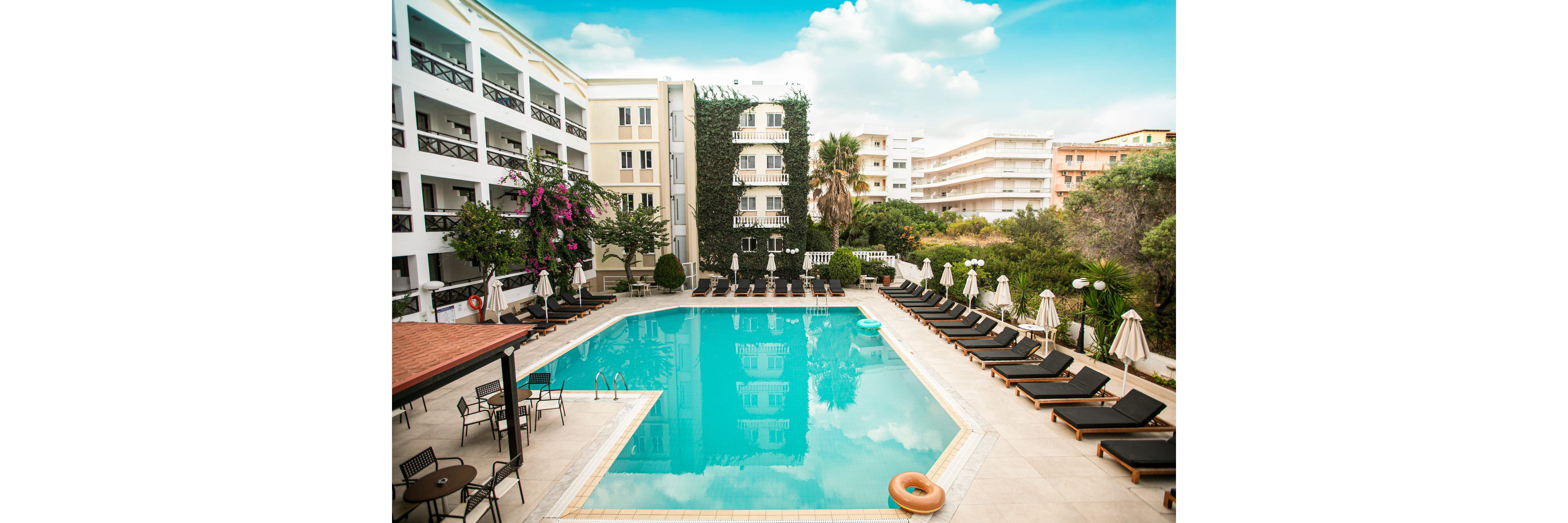 A large rectangular swimming pool sits at the center of a modern hotel courtyard. Rows of black lounge chairs and beige umbrellas line both sides of the pool, creating a resort-like atmosphere. The surrounding multi-story buildings feature balconies, greenery, and flowering plants, with palm trees adding a tropical touch. The sky is bright with scattered clouds, suggesting a warm and inviting day for relaxation by the water.