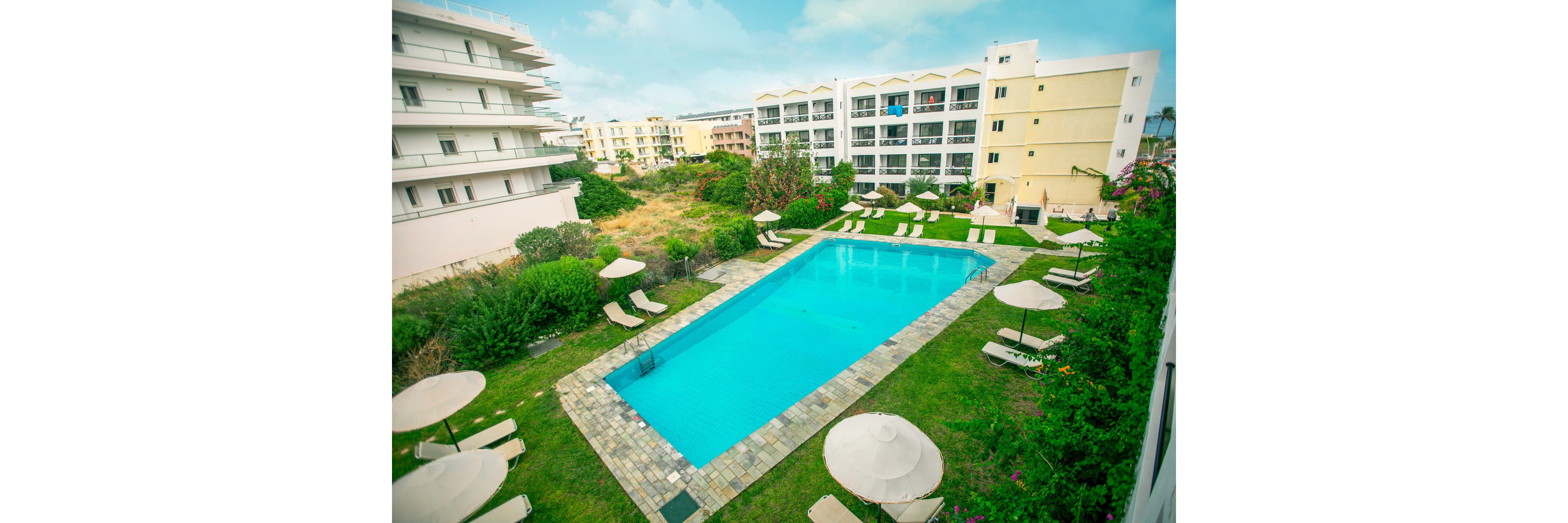 An outdoor rectangular swimming pool is centered between modern hotel buildings. Numerous sun loungers and umbrellas are arranged neatly around the pool on a manicured lawn. The scene suggests a resort setting with landscaped greenery and a relaxed vacation atmosphere. Bright daylight and a partly cloudy sky create a fresh, inviting mood.