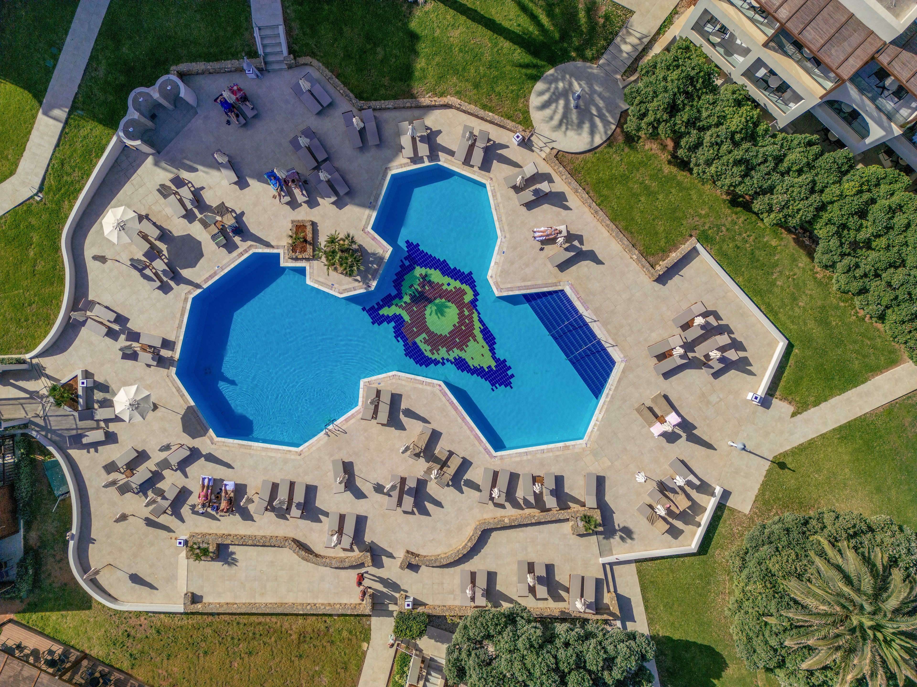A large geometric swimming pool is photographed from above in a resort setting. The pool features a colorful mosaic design at its center and is surrounded by numerous lounge chairs and umbrellas. Green lawns, trees, and landscaped gardens border the paved pool deck. A few people relax on sunbeds, emphasizing a calm vacation atmosphere.