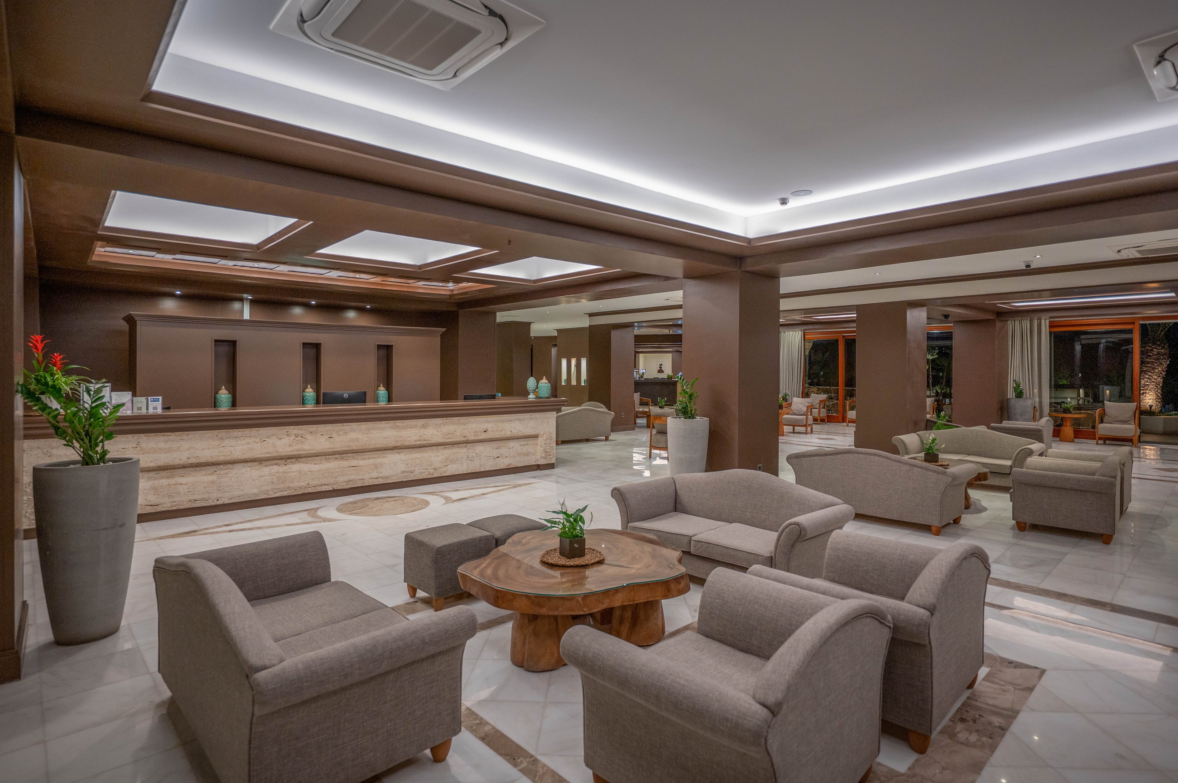Spacious hotel lobby interior featuring a central seating area with cushioned armchairs around a wooden coffee table. The reception desk extends along the back wall, illuminated by recessed ceiling lights and indirect LED lighting. Large potted plants add greenery to the neutral brown and beige color palette. Polished tile floors and contemporary furnishings create a clean, upscale atmosphere.