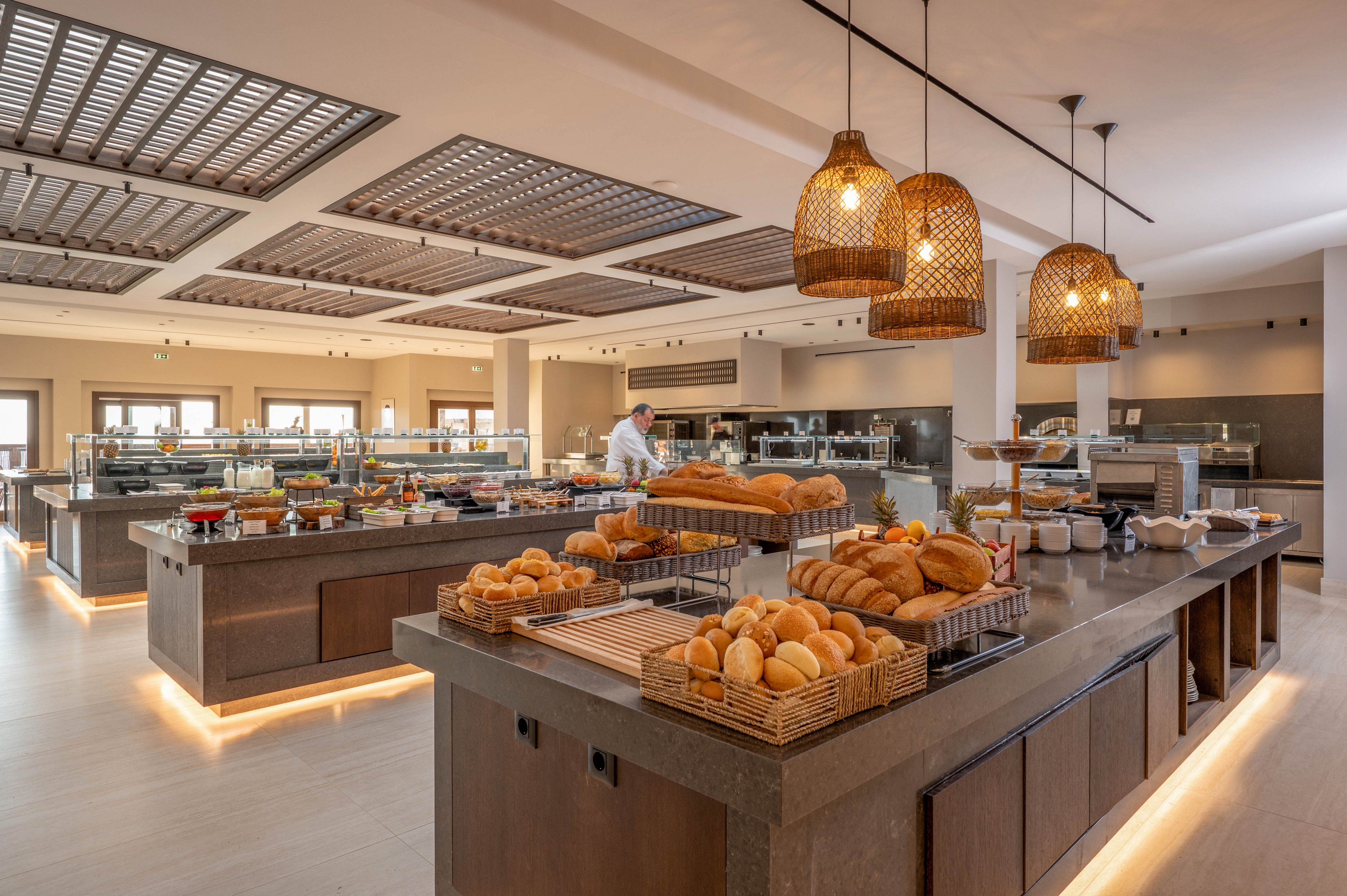 A spacious self-service buffet area showcases an assortment of fresh bread and rolls arranged in baskets on a central island. In the background, a chef works behind the counters, preparing additional dishes in a clean, contemporary dining space. Warm pendant lights and under-counter lighting create a cozy, inviting atmosphere. The neutral color palette and organized layout emphasize a modern, upscale hotel or resort restaurant setting.