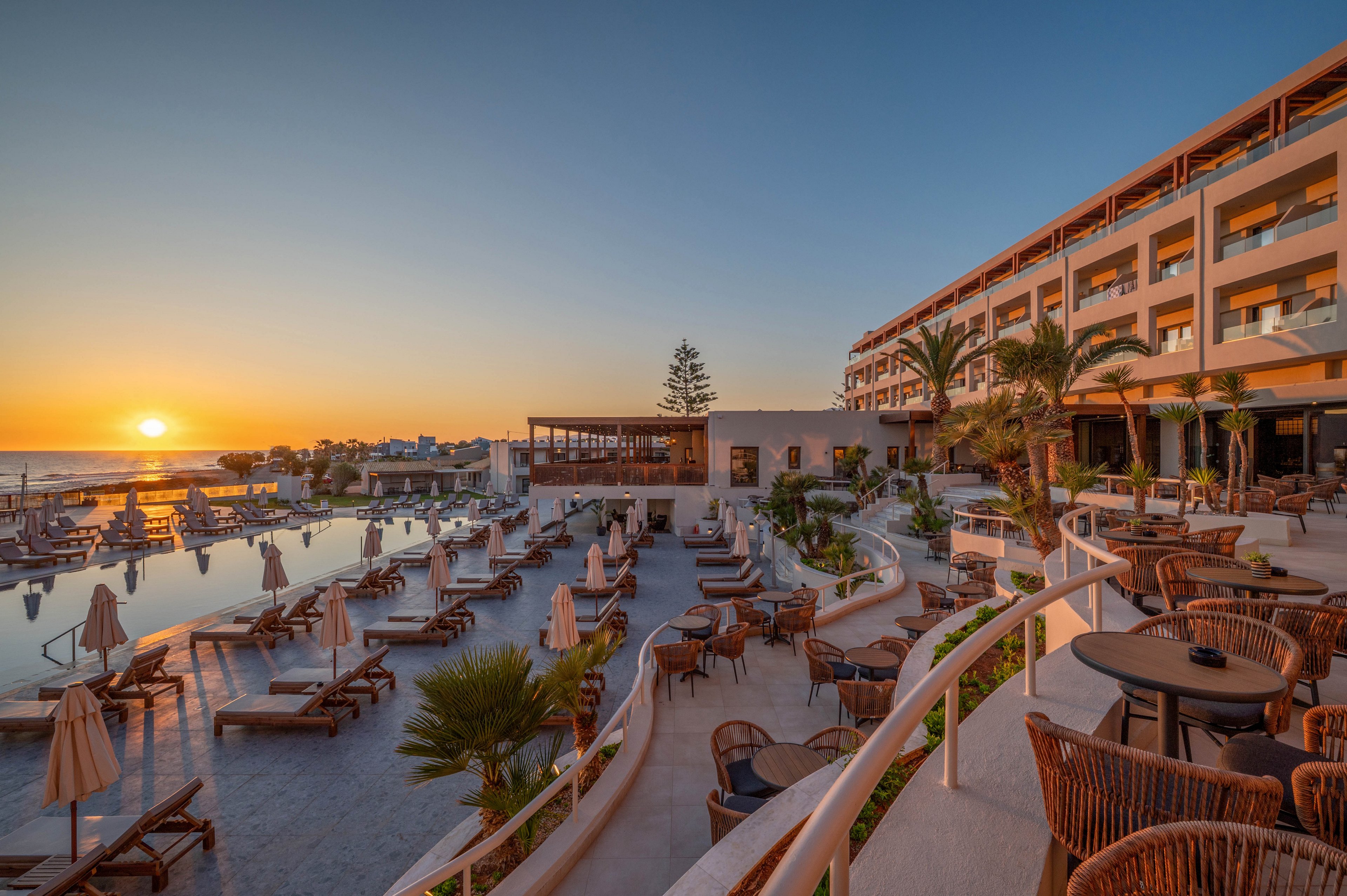 A luxurious seaside resort features an expansive pool area lined with sun loungers and umbrellas at sunset. The multi-story hotel building overlooks the water, with balconies facing the ocean horizon. Outdoor seating areas with wicker chairs and tables create an inviting atmosphere for guests to relax. Warm golden light from the setting sun reflects on the pool surface, enhancing the tranquil vacation mood.
