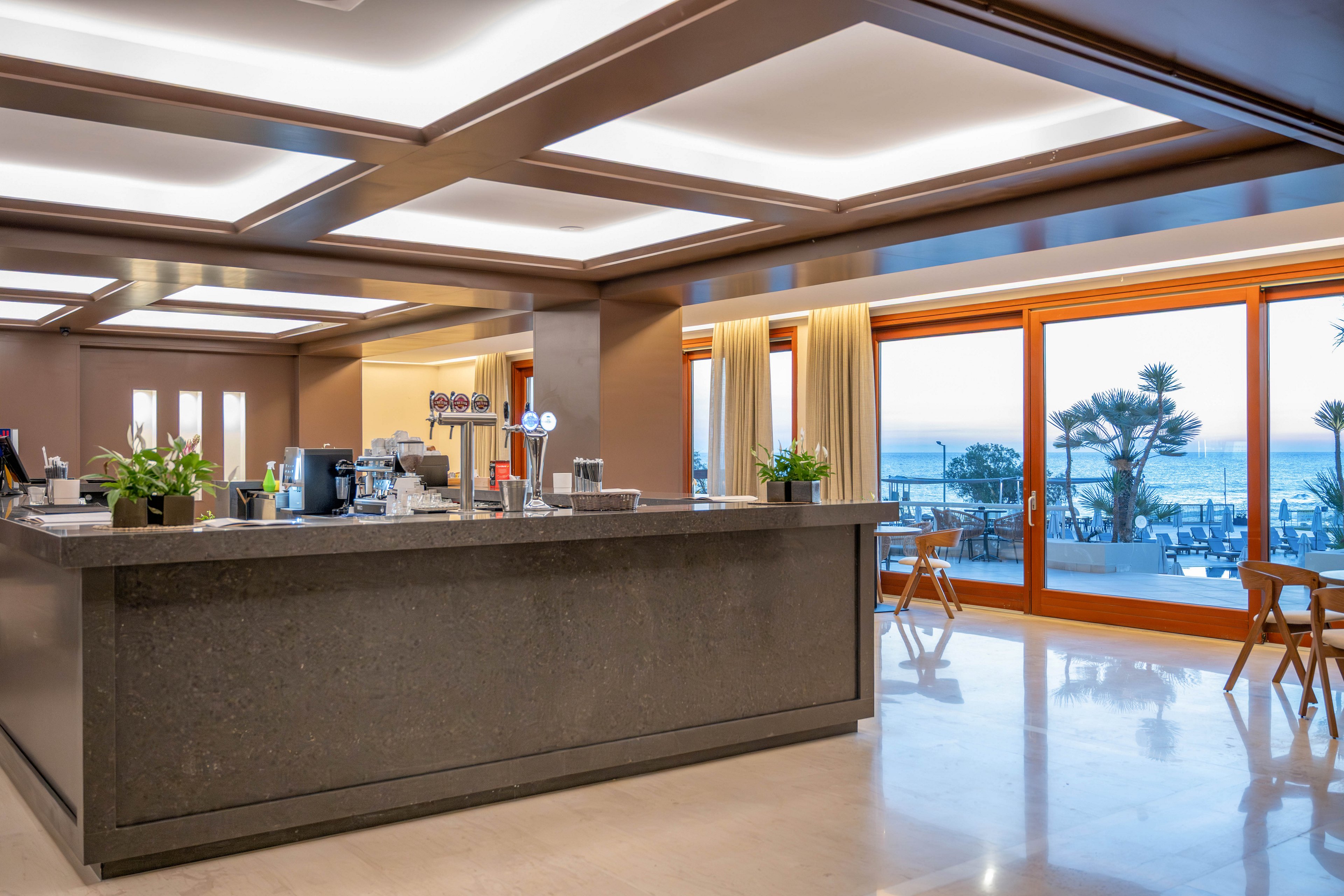 A sleek, modern hotel bar counter stands in the foreground of a spacious interior. Large floor-to-ceiling windows reveal an outdoor terrace with seating and a scenic ocean view at sunset. Warm ceiling lights and wooden accents create a cozy, upscale atmosphere. Potted plants and bar equipment add detail to the contemporary hospitality setting.