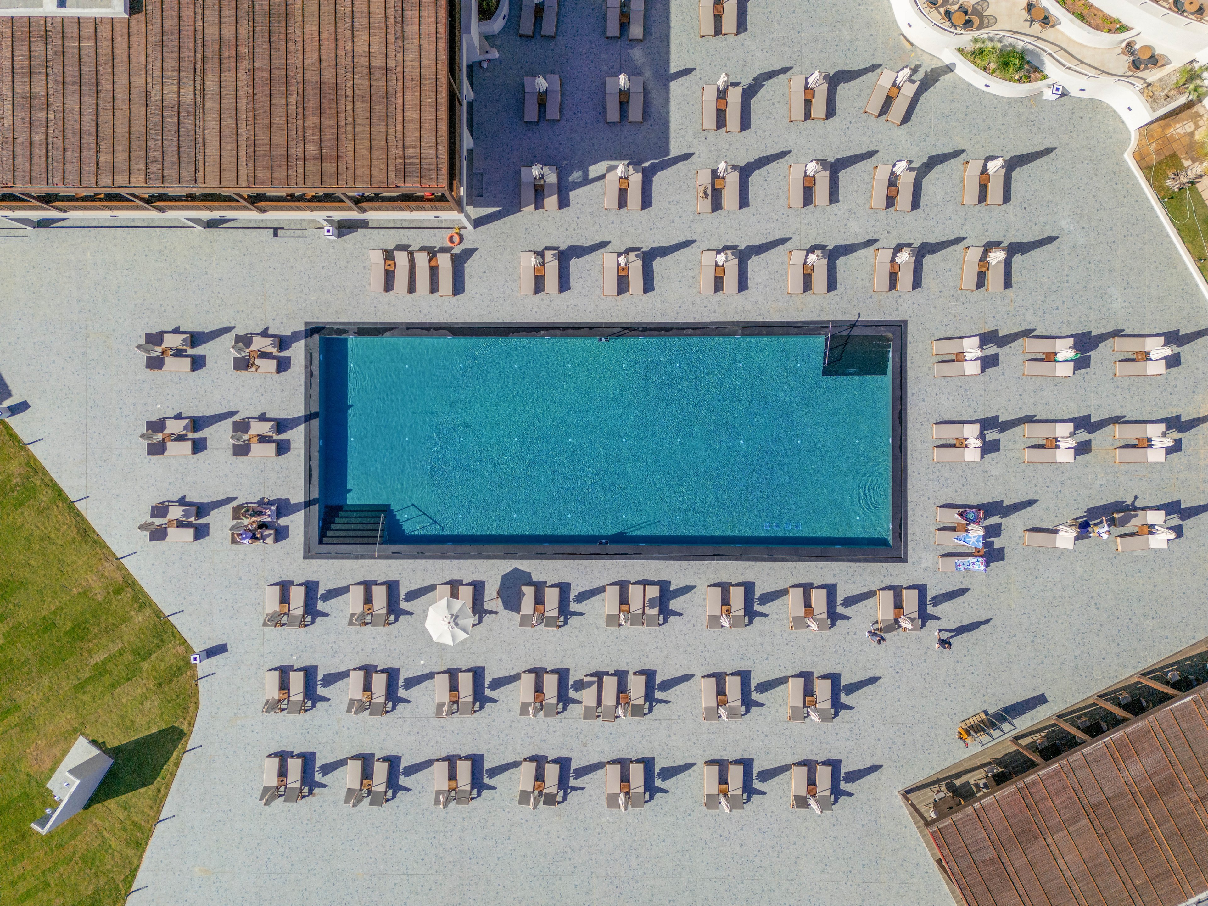 An aerial view shows a large rectangular swimming pool surrounded by neatly arranged lounge chairs. The pool is set within a resort-style terrace with buildings framing two sides of the scene. Sun loungers and umbrellas create a repeating geometric pattern around the water. A few people can be seen relaxing near the pool, emphasizing a calm vacation atmosphere. The overall setting suggests a sunny, organized, and upscale outdoor leisure area.
