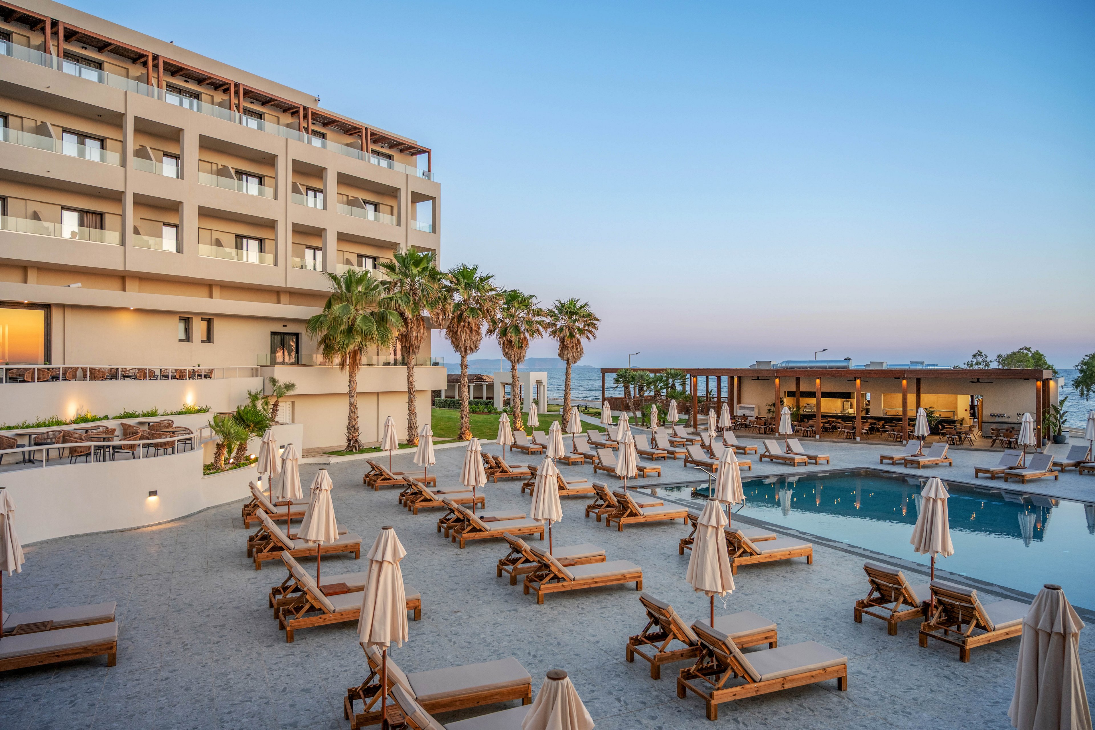 A modern multi-story seaside resort overlooks a tranquil outdoor pool area. Numerous wooden sun loungers with beige umbrellas are neatly arranged around the pool on a stone terrace. Palm trees and a low pavilion structure frame the scene, with the ocean visible in the background. The soft evening sky creates a calm, relaxing vacation atmosphere.