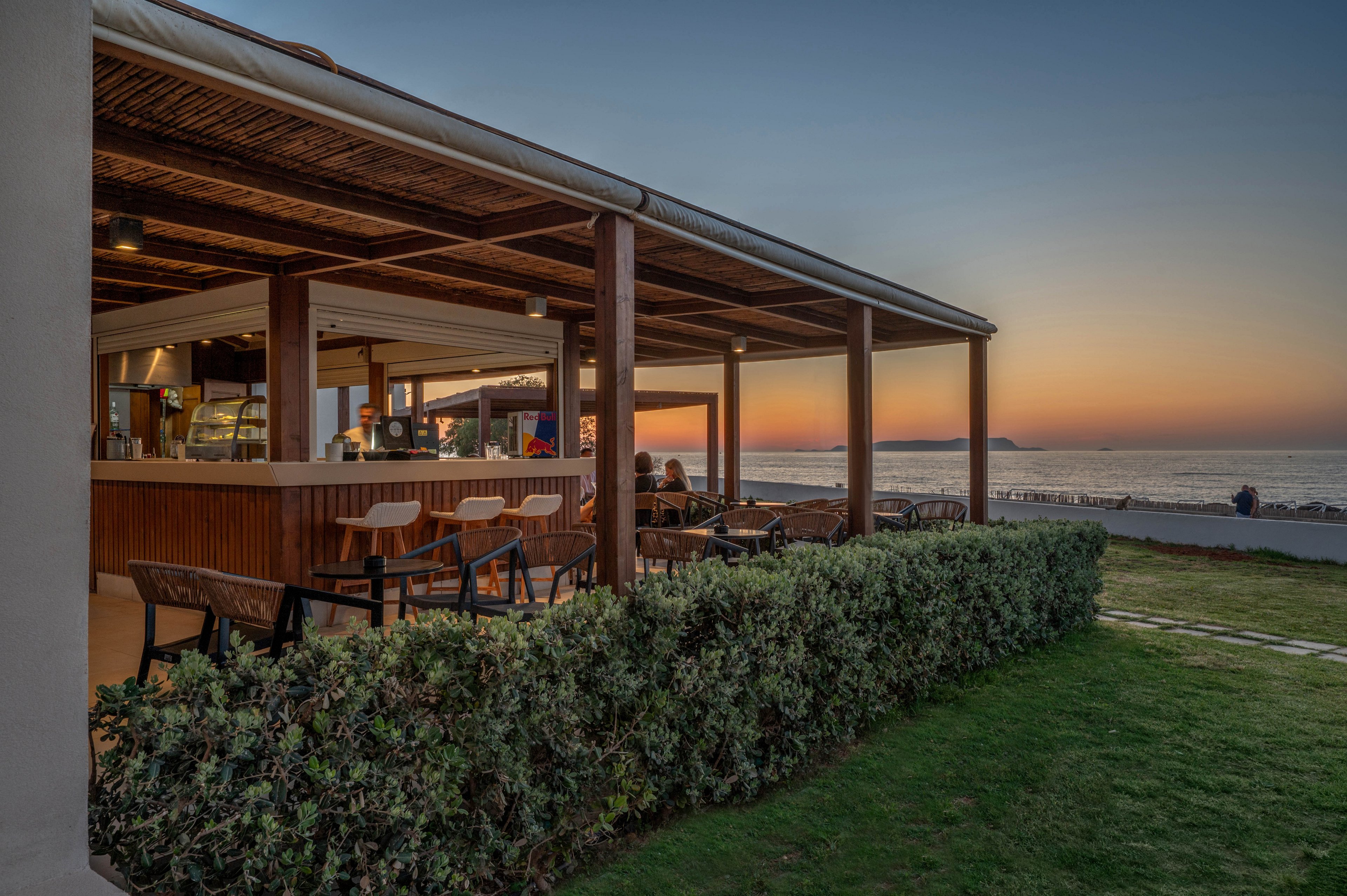 An open-air beachfront bar with wooden beams and modern seating overlooks the sea at sunset. A few people sit inside the bar area while the terrace remains mostly empty. Lush green hedges and a manicured lawn frame the structure, leading toward the shoreline. The sky glows with warm orange and blue tones as the sun sets over the calm water.
