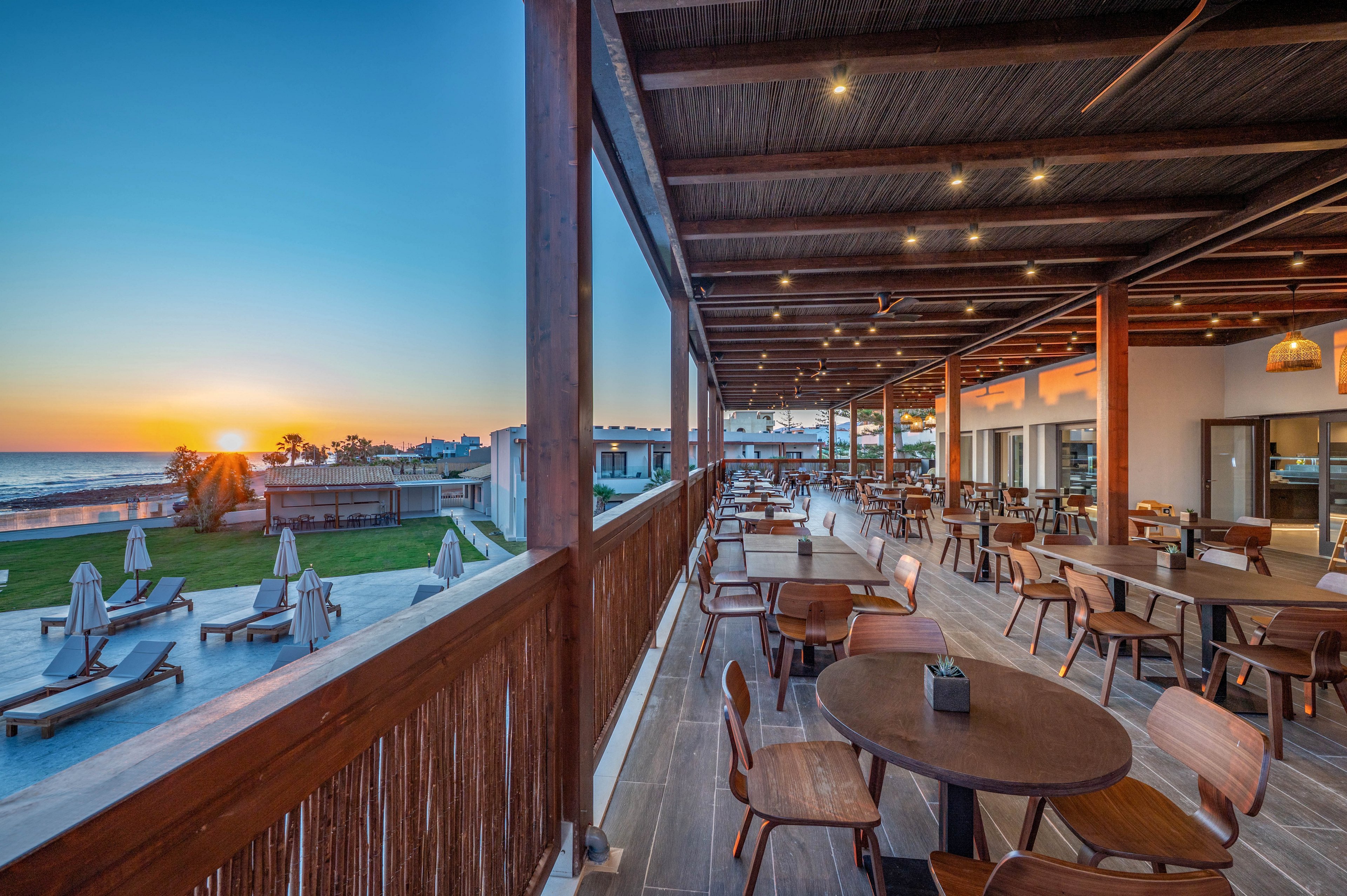 An open-air restaurant terrace overlooks a coastal resort at sunset. Wooden tables and chairs are neatly arranged under a covered patio with warm ceiling lights. In the background, sun loungers and umbrellas face the calm sea and glowing horizon. The scene feels tranquil and inviting, ideal for relaxed outdoor dining by the water.