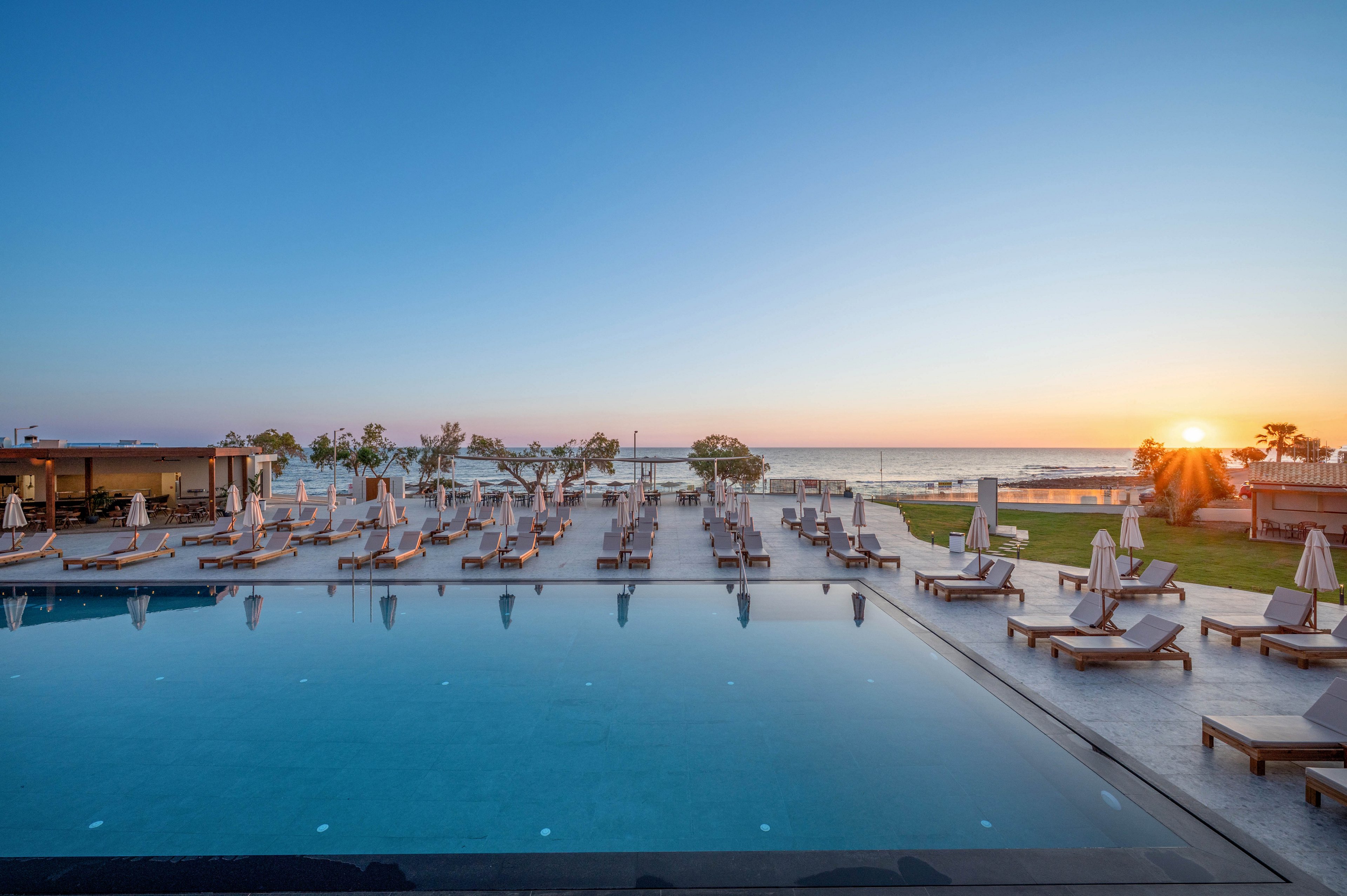 A large infinity-style swimming pool sits at the center of a modern beachfront resort. Numerous sun loungers and umbrellas are neatly arranged around the pool, facing the sea. The sun is setting over the horizon, casting warm light across the calm water and clear sky. A pool bar and landscaped lawn area complete the tranquil holiday setting.