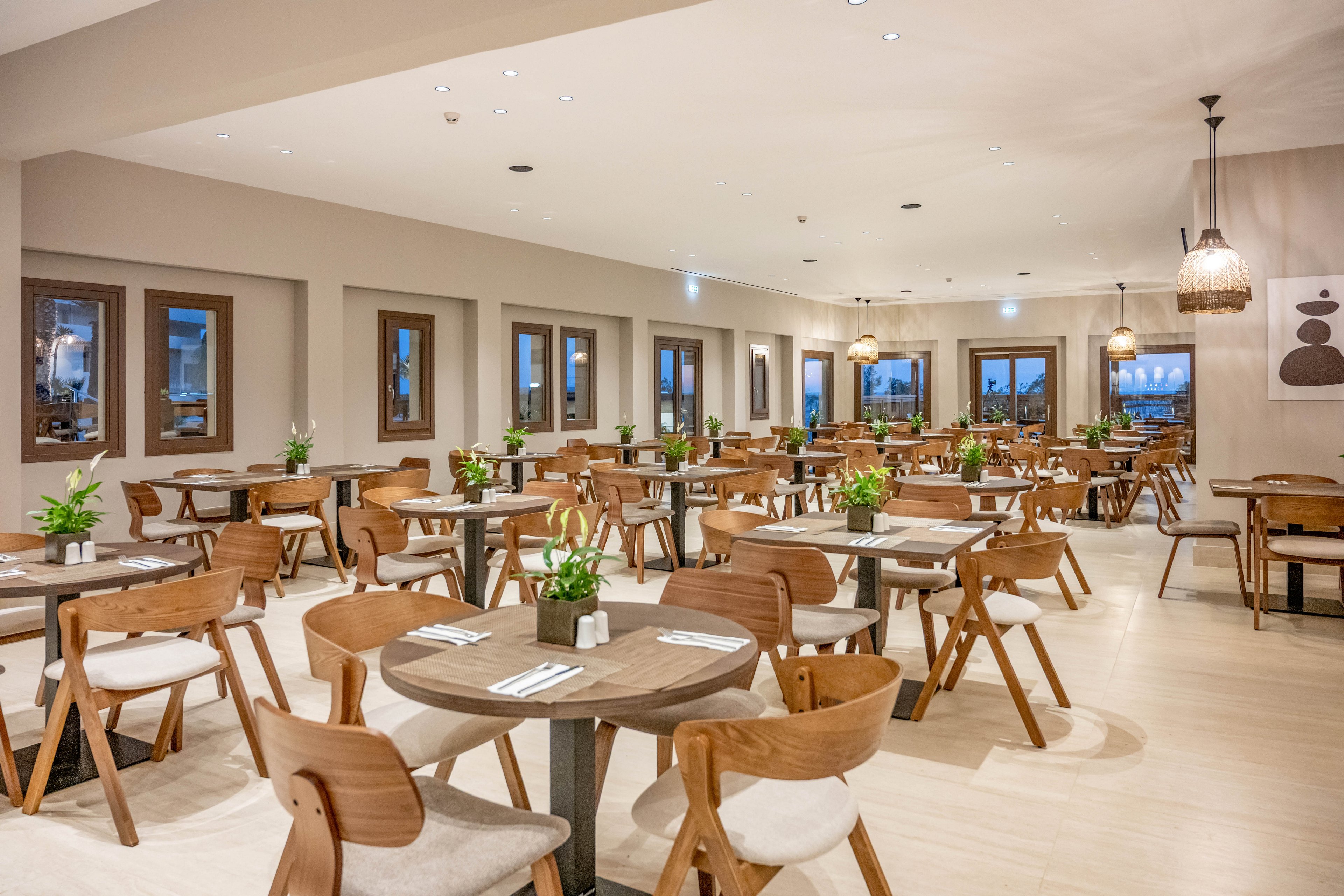 Spacious contemporary restaurant dining room with neatly arranged wooden tables and chairs. The setting is bright and clean, featuring neutral walls, light flooring, and large windows along one side. Small green plants decorate each table, and simple artwork and pendant lights add a warm, modern touch. The room is empty of people, emphasizing order, symmetry, and a calm atmosphere.