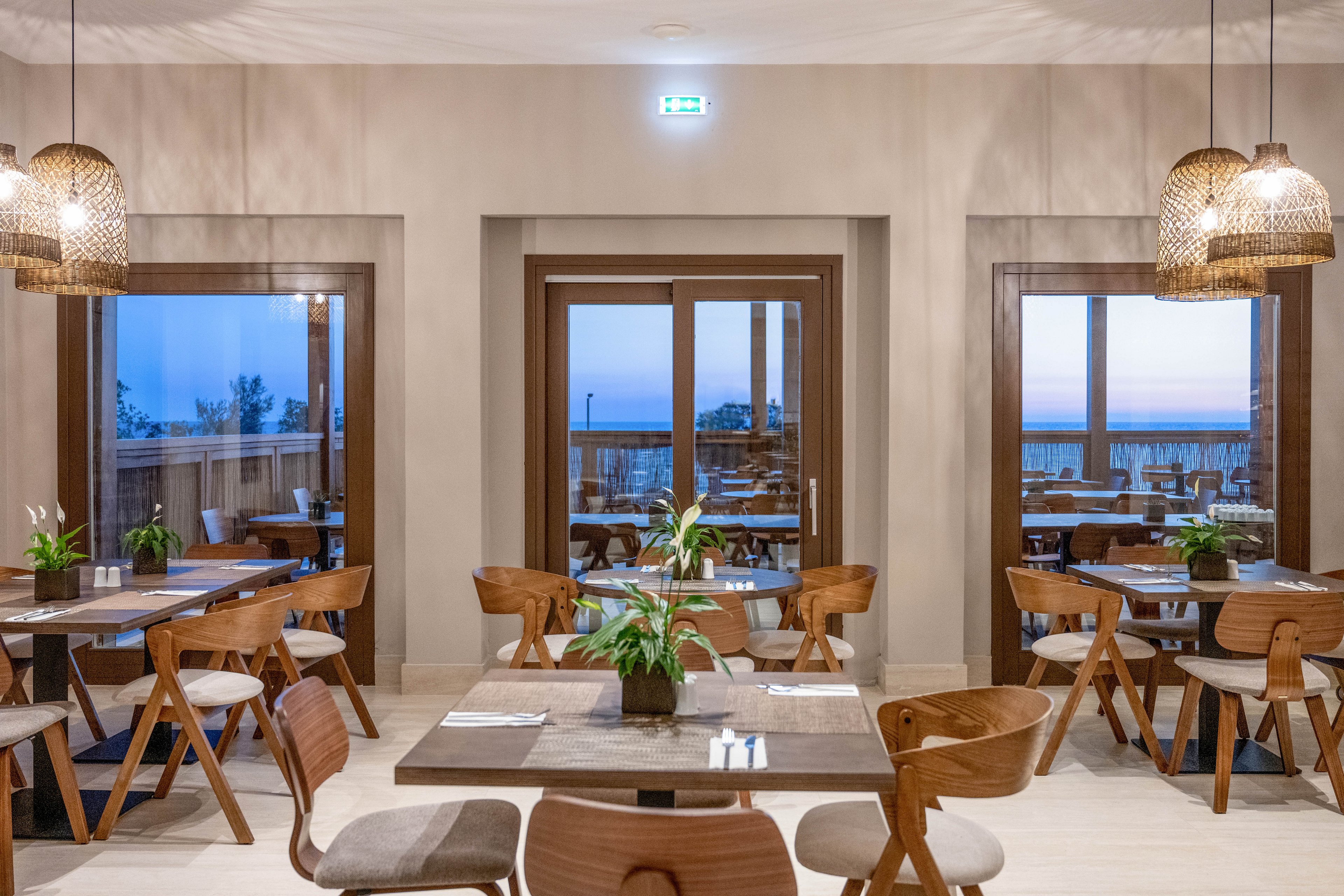 Contemporary restaurant dining area with wooden tables and chairs is shown in soft evening light. Large windows and glass doors reveal a calm seaside view and blue twilight sky outside. Pendant wicker lamps hang from the ceiling, casting a warm glow over the neutral-toned space. Green potted plants on the tables add a fresh, natural accent to the minimalist decor.