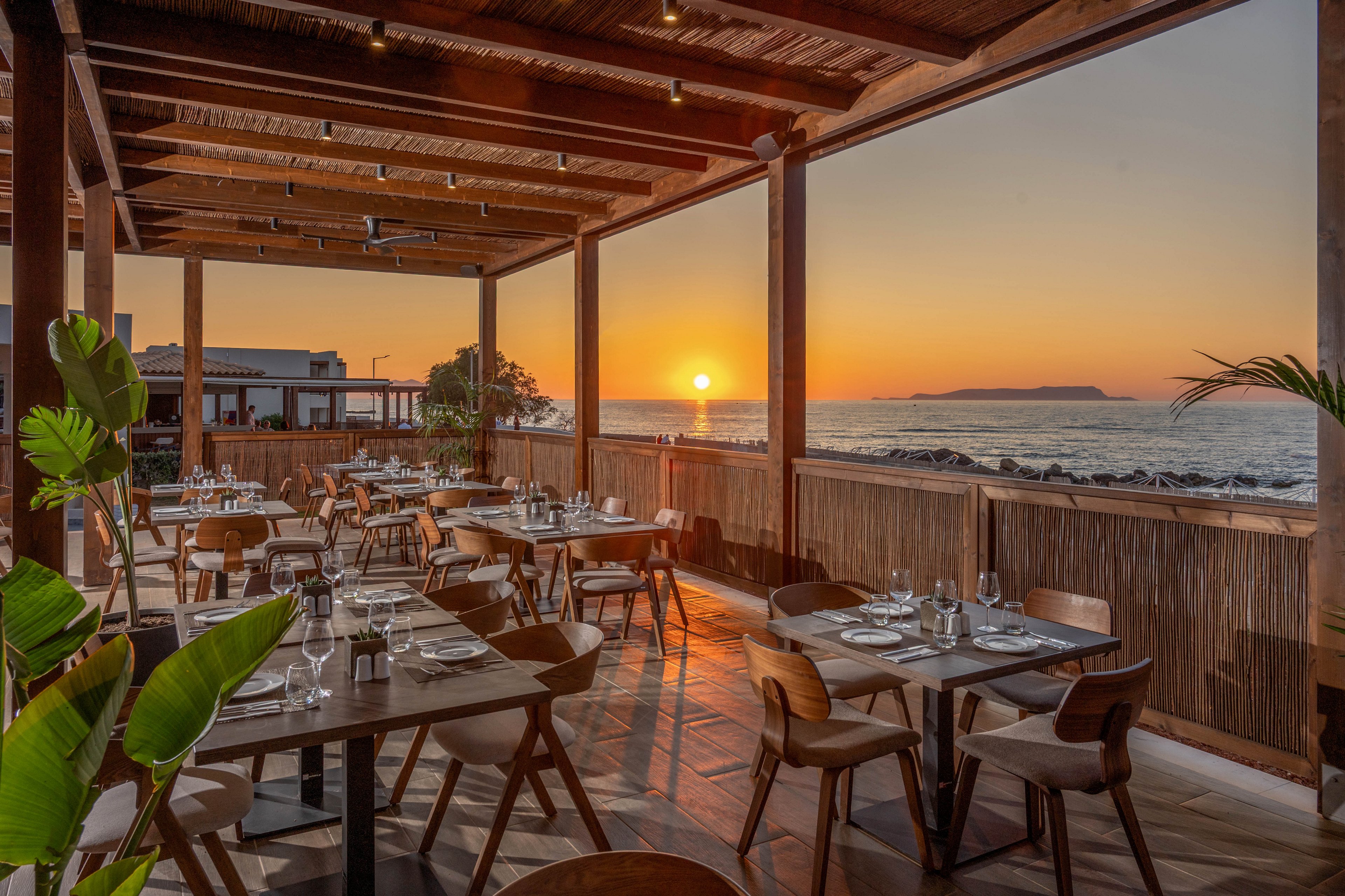 Open-air restaurant tables are set neatly on a wooden terrace overlooking the sea. The sun is setting on the horizon, casting warm golden light across the water and interior. Empty chairs and arranged glassware suggest the space is ready for evening diners. Natural materials, plants, and soft colors create a relaxed coastal atmosphere.