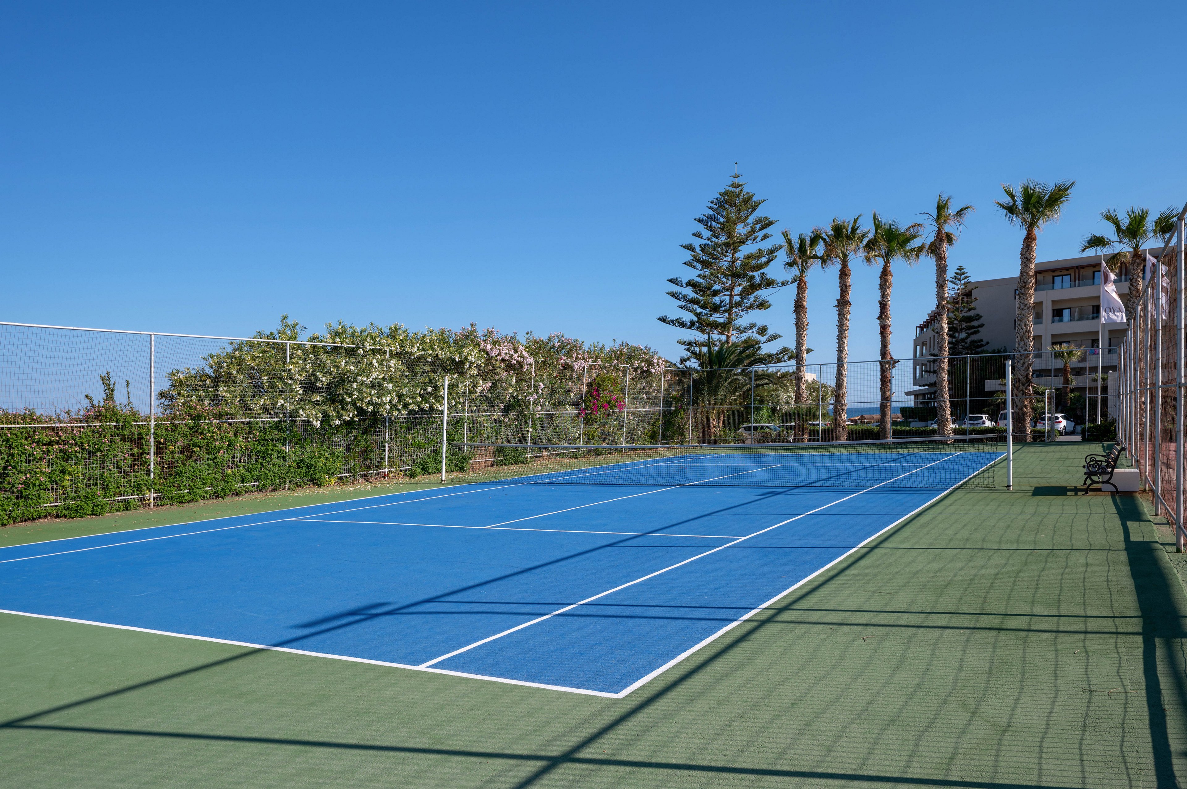 A bright blue tennis court sits empty under a clear blue sky. The court is surrounded by green fencing, lush hedges, and tall palm trees. In the background, a multi-story building suggests a coastal hotel or resort setting. Strong sunlight and sharp shadows create a clean, summery atmosphere.