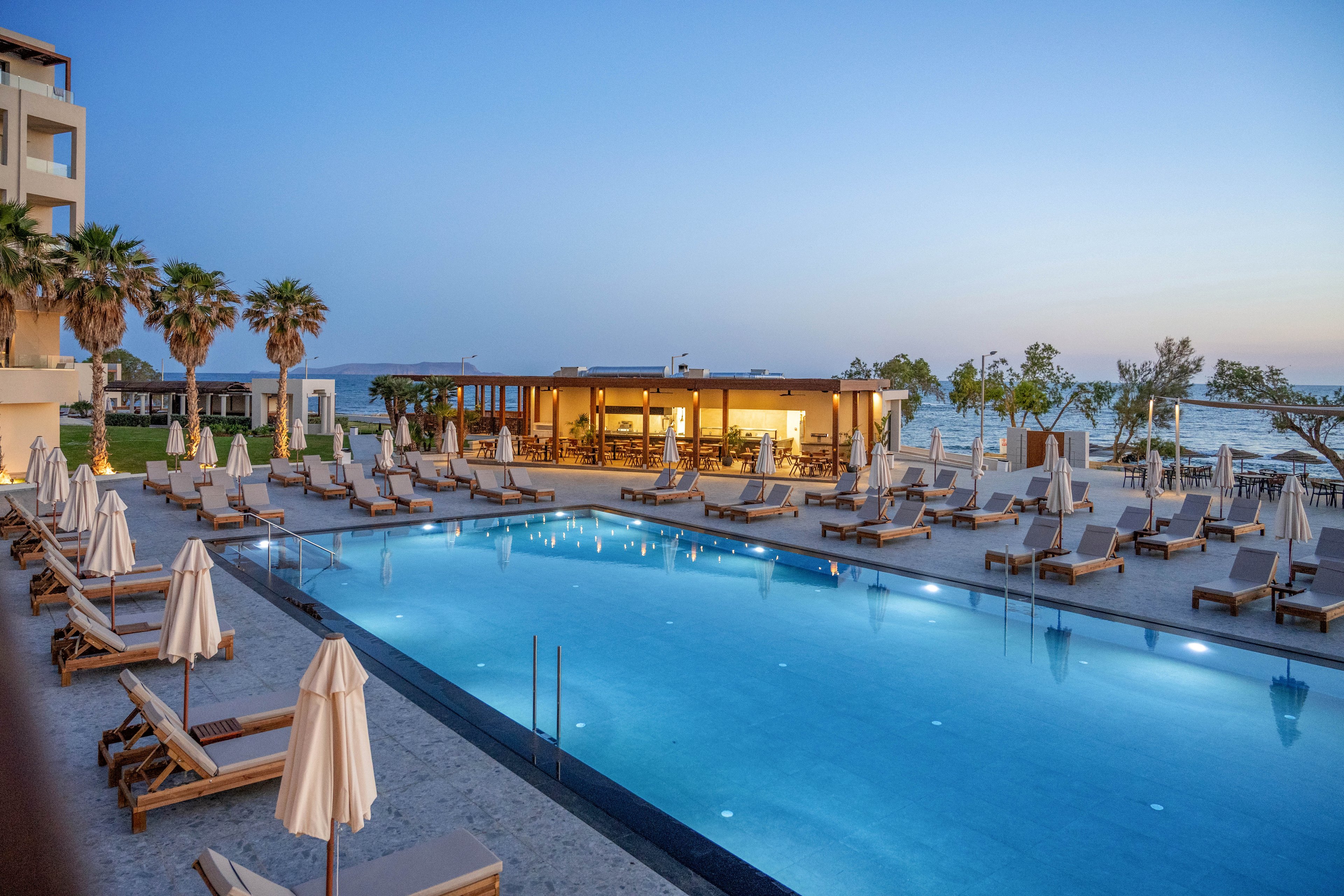 A large illuminated swimming pool is surrounded by neatly arranged sun loungers and closed umbrellas at a seaside resort. The scene is captured at dusk with a soft blue sky and calm ambient lighting. Palm trees and low buildings frame the pool area, leading the eye toward the ocean in the background. The tranquil atmosphere suggests a relaxing vacation destination by the water.