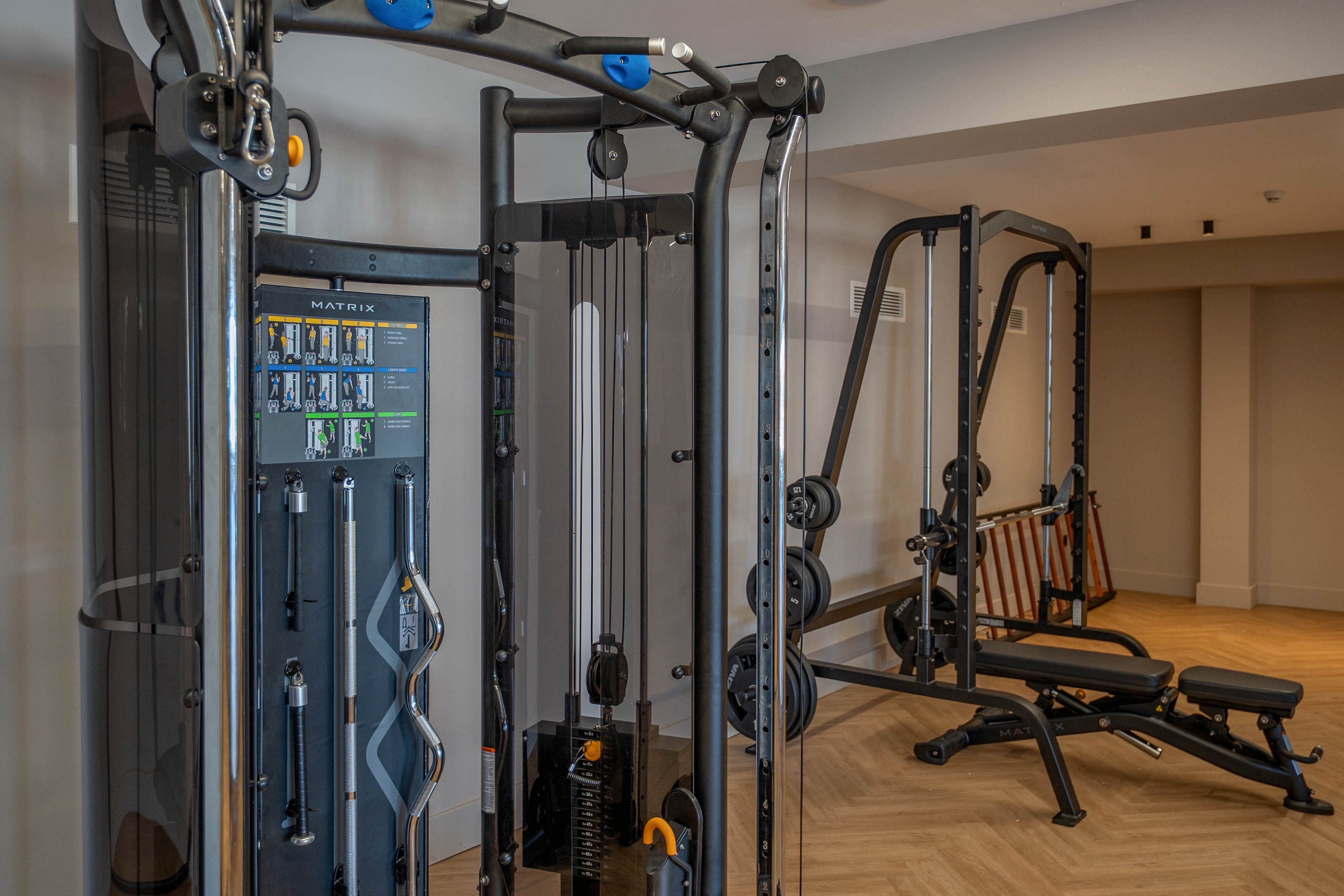 A compact fitness room features modern multi-station strength training equipment. The machines are arranged on a light wood herringbone floor under a sleek linear ceiling light. Neutral walls and minimal decor create a clean, contemporary workout environment. The space appears well-maintained and designed for functional training and weight exercises.