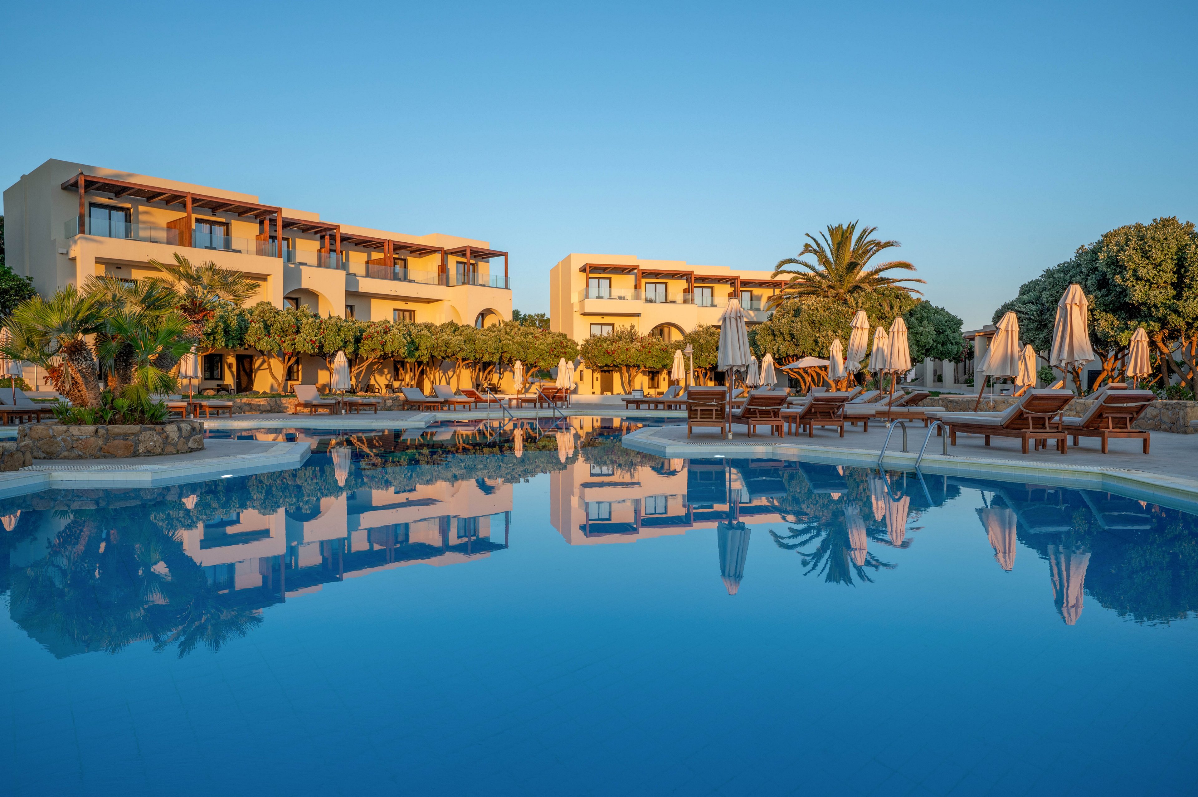 A serene resort features a large outdoor swimming pool reflecting the surrounding buildings and clear blue sky. Sun loungers and closed umbrellas line the poolside, suggesting a quiet moment before or after peak activity. Warm sunlight bathes the modern, low-rise hotel structures and lush greenery, creating a calm and inviting atmosphere. The scene evokes a peaceful vacation setting ideal for relaxation and leisure travel.