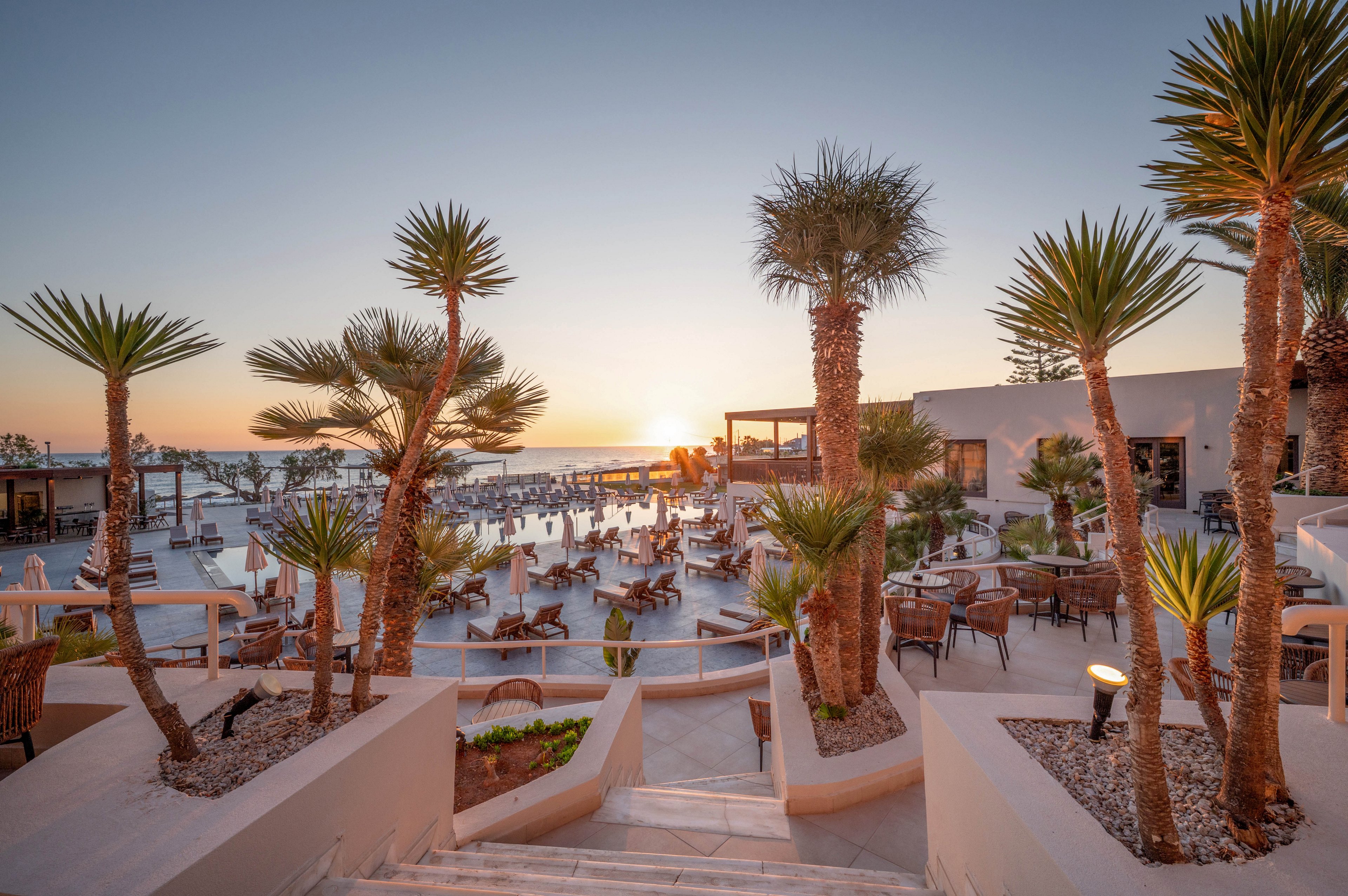A contemporary seaside resort is captured at sunset, with warm light reflecting off a central swimming pool. Palm trees and desert-style plants line the stepped pathways leading down to the pool area. Numerous lounge chairs and outdoor seating are arranged around the water, facing the ocean horizon. The scene conveys a relaxed vacation atmosphere with soft pastel sky tones and calm coastal surroundings.