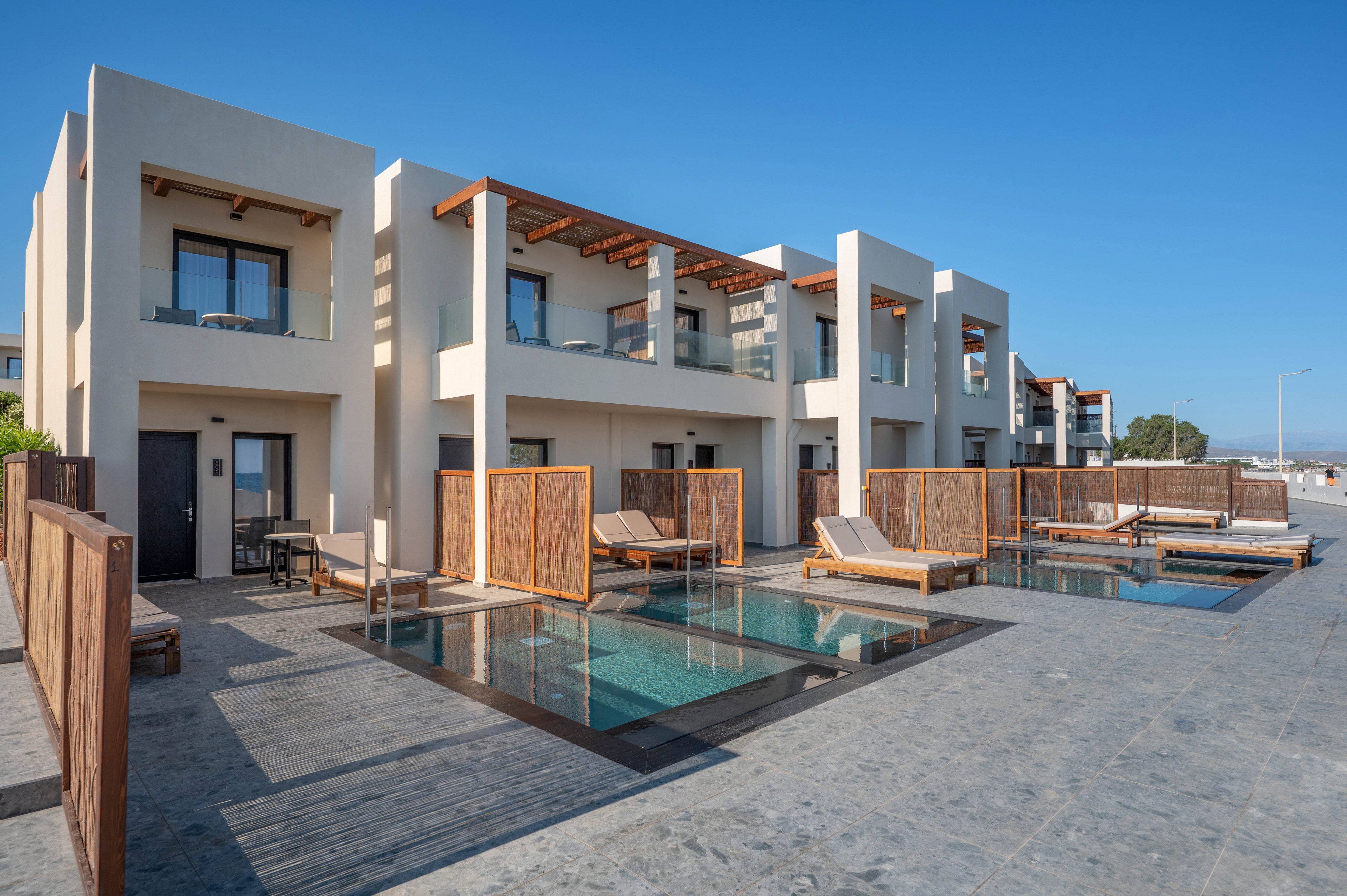 Contemporary two-story villas feature clean, cubic architecture and private plunge pools in front of each unit. Sun loungers and wooden privacy screens create an intimate resort atmosphere. The scene is set under a clear blue sky, suggesting a warm coastal or island destination. The overall look is minimalist and luxurious, emphasizing outdoor relaxation and modern design.