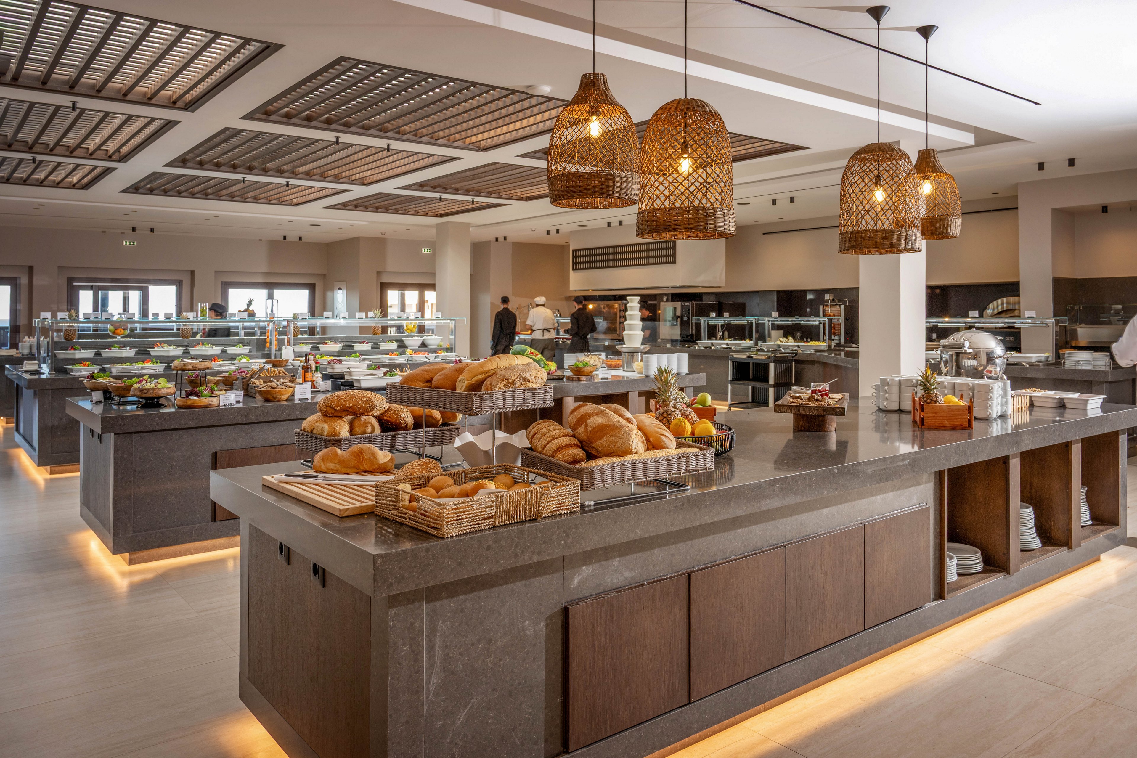 A spacious self-service buffet area showcases an assortment of fresh breads and pastries on a central stone counter. Wicker baskets hold loaves, rolls, and croissants, while additional counters in the background display salads, fruits, and other dishes. Warm pendant lights and under-counter lighting create a cozy, contemporary atmosphere. A few people are visible in the distance, adding a subtle sense of activity to the otherwise calm dining space.