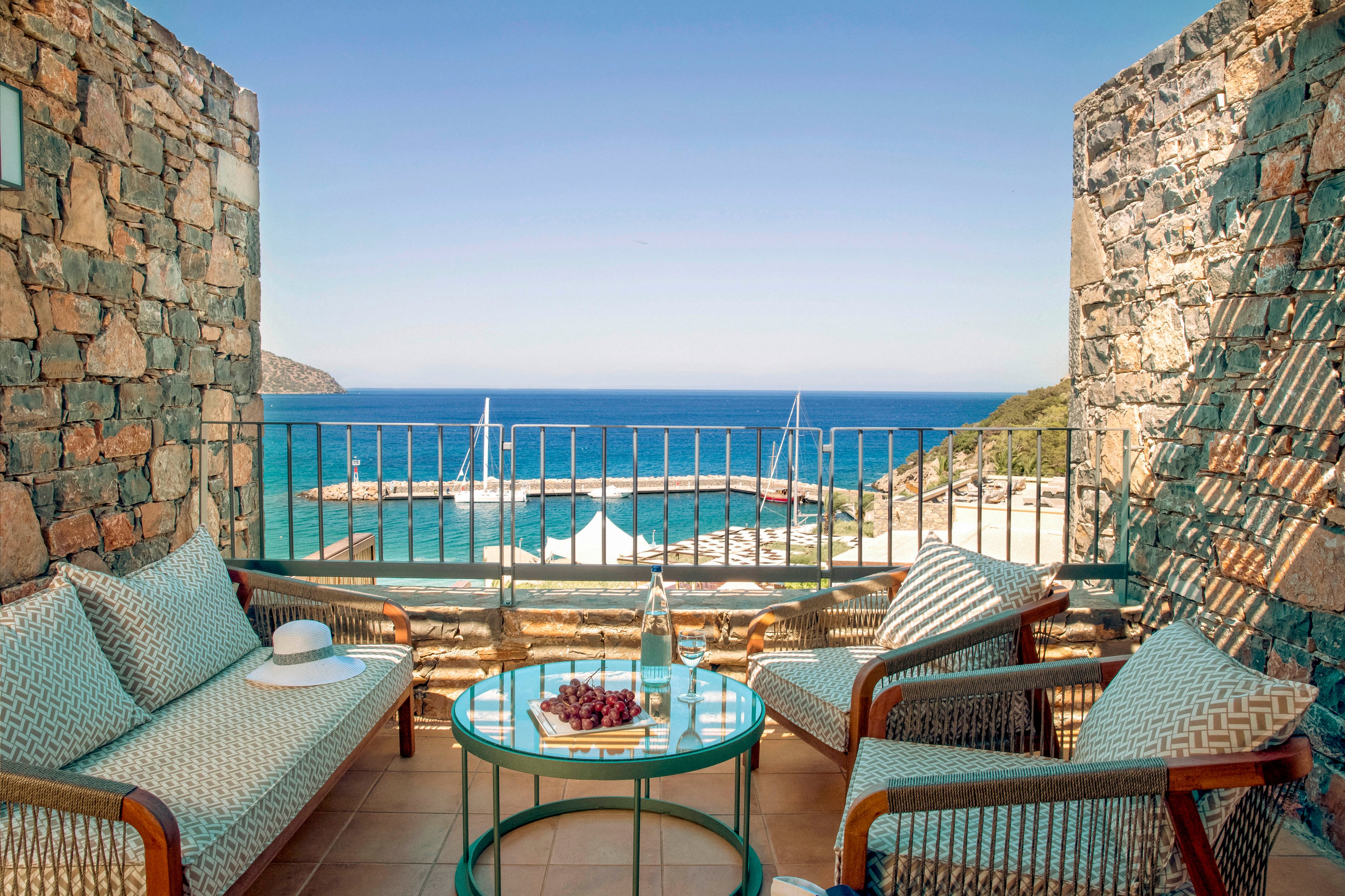 An inviting terrace seating area opens to a scenic view of a bright blue sea and small marina. Cushioned chairs and a sofa with patterned fabric surround a round glass table holding a bottle, glasses, and a plate of grapes. Stone walls frame the balcony, while a metal railing looks out toward sailboats and a sandy shoreline. The warm sunlight and calm water create a relaxed, luxurious vacation atmosphere.