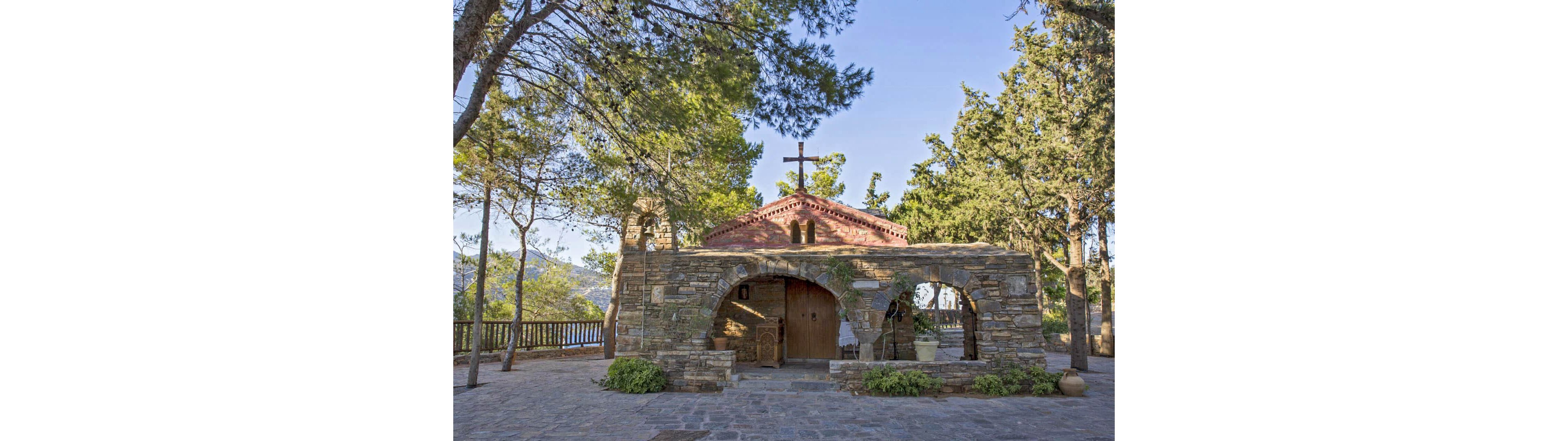 A small stone chapel stands in a peaceful hillside setting surrounded by tall pine and cypress trees. The building features arched entrances, a rustic stone façade, and a red-tiled roof topped with a simple cross. Sunlight filters through the trees, casting soft shadows on the cobblestone courtyard in front of the chapel. A wooden fence and distant hills are visible in the background, enhancing the tranquil, rural atmosphere.